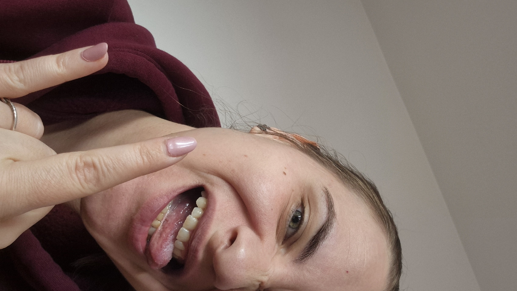 A woman is pictured from a low angle, tilted to the right. She is wearing a maroon hoodie and has a ring on her finger. Her tongue is sticking out, and she is making a gesture with her fingers, pointing towards herself. Her fingernails are manicured with a light pink color. An adhesive bandage is visible on her forehead near her hairline. The background appears to be a plain wall. No information is available to determine whether the picture was taken in Sofia, Bulgaria.