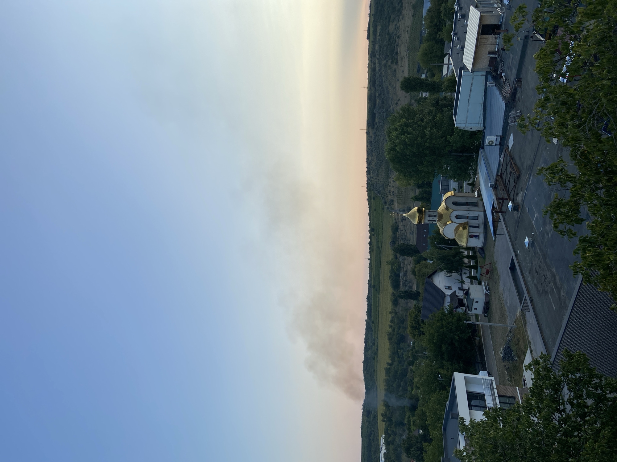 The image shows a view of a town with buildings, trees, and a hill in the background. A plume of smoke is visible rising from behind a line of trees on the hill. The sky is clear with a gradient of blue to pale orange, indicating either early morning or late evening. In the foreground, there are several buildings with flat roofs. A distinctive golden-domed church is visible among the buildings, and a modern white building with dark windows is also present. Trees are scattered throughout the scene, some with dark green foliage. The overall impression is of a rural or semi-urban environment experiencing an event that is producing smoke in the distance.