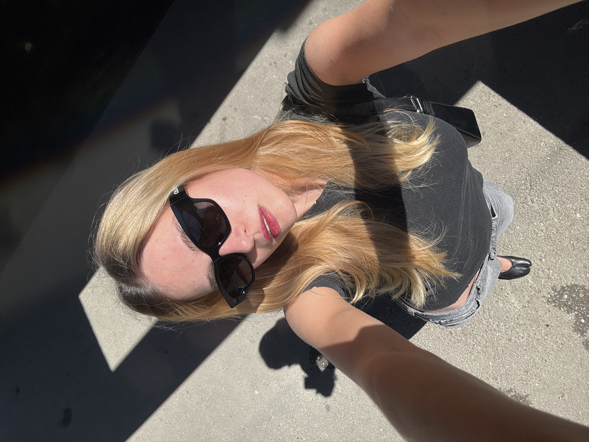 A high-angle, selfie-style photograph shows a woman with long, blonde hair and sunglasses, standing on a paved surface in direct sunlight. She is wearing a dark top and light-colored jeans, secured with a black belt. Her visible footwear consists of a single black shoe, and she is holding a black object, possibly a phone, in her right hand. Harsh shadows are cast on the ground, suggesting a bright, sunny day in an urban environment. The specific location in the city of Unknown, Unknown cannot be determined.