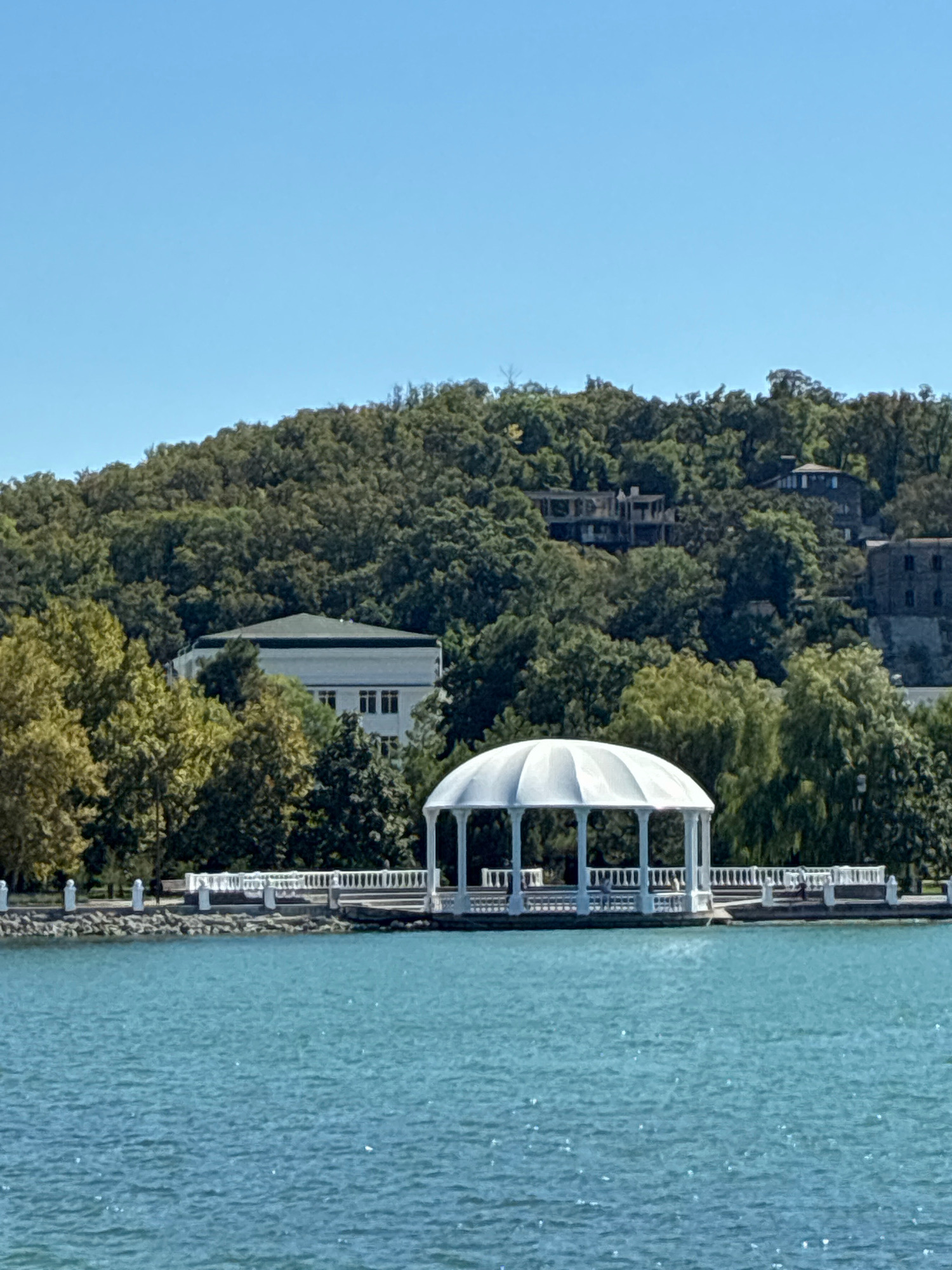 The image depicts a waterfront scene in Abrau-Dyurso, Russia, under a clear blue sky. In the foreground, a tranquil, turquoise body of water occupies the bottom half of the frame. Along the water's edge, a white, ornate gazebo with a domed roof stands prominently, supported by pillars. A white, decorative railing lines the promenade behind the gazebo, separating it from the land.
Beyond the waterfront, a lush, green hillside is densely covered with trees. Several buildings are visible nestled amongst the foliage on the slope. One building, partially obscured by trees, appears to be a white, multi-story structure with a dark roof. Further up the hill, other buildings, some with darker facades, are discernible. The overall impression is one of a serene, scenic location, likely a park or resort area. The image appears to be taken during daylight hours, suggesting a pleasant weather day. No people are clearly visible in the image.