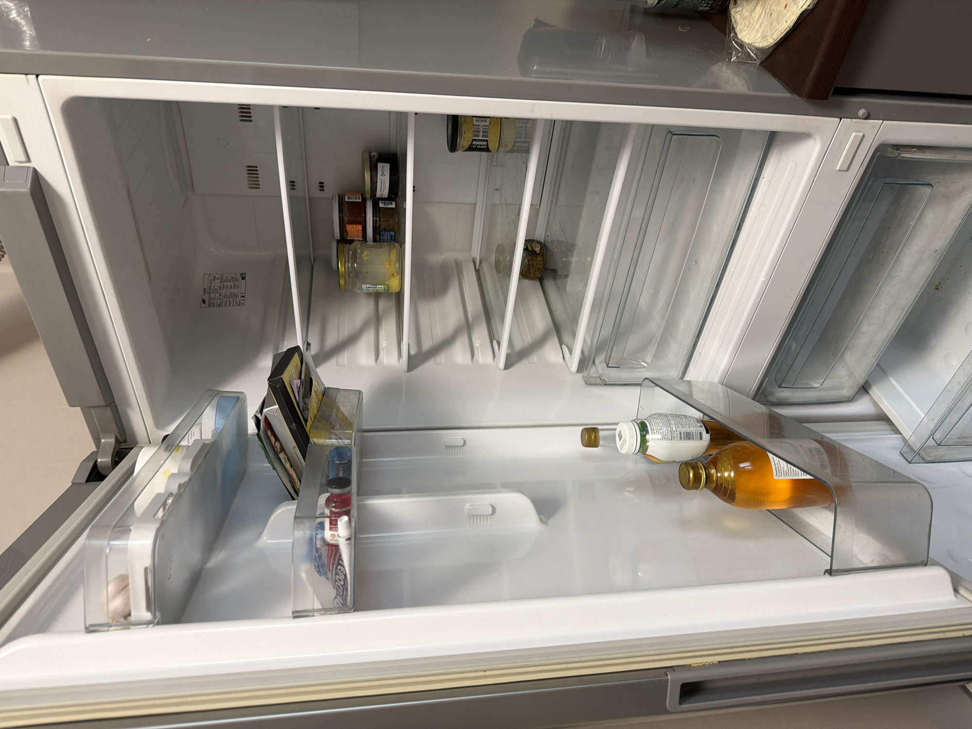 The image shows the interior of a refrigerator. The refrigerator appears to be mostly empty, containing jars and bottles. There are two bottles in a rack, one with a clear liquid and the other with an amber-colored liquid. The scene is in a neutral, indoor environment, possibly a home kitchen. There is no information to connect the image to the city of Saint Petersburg, Russia.
