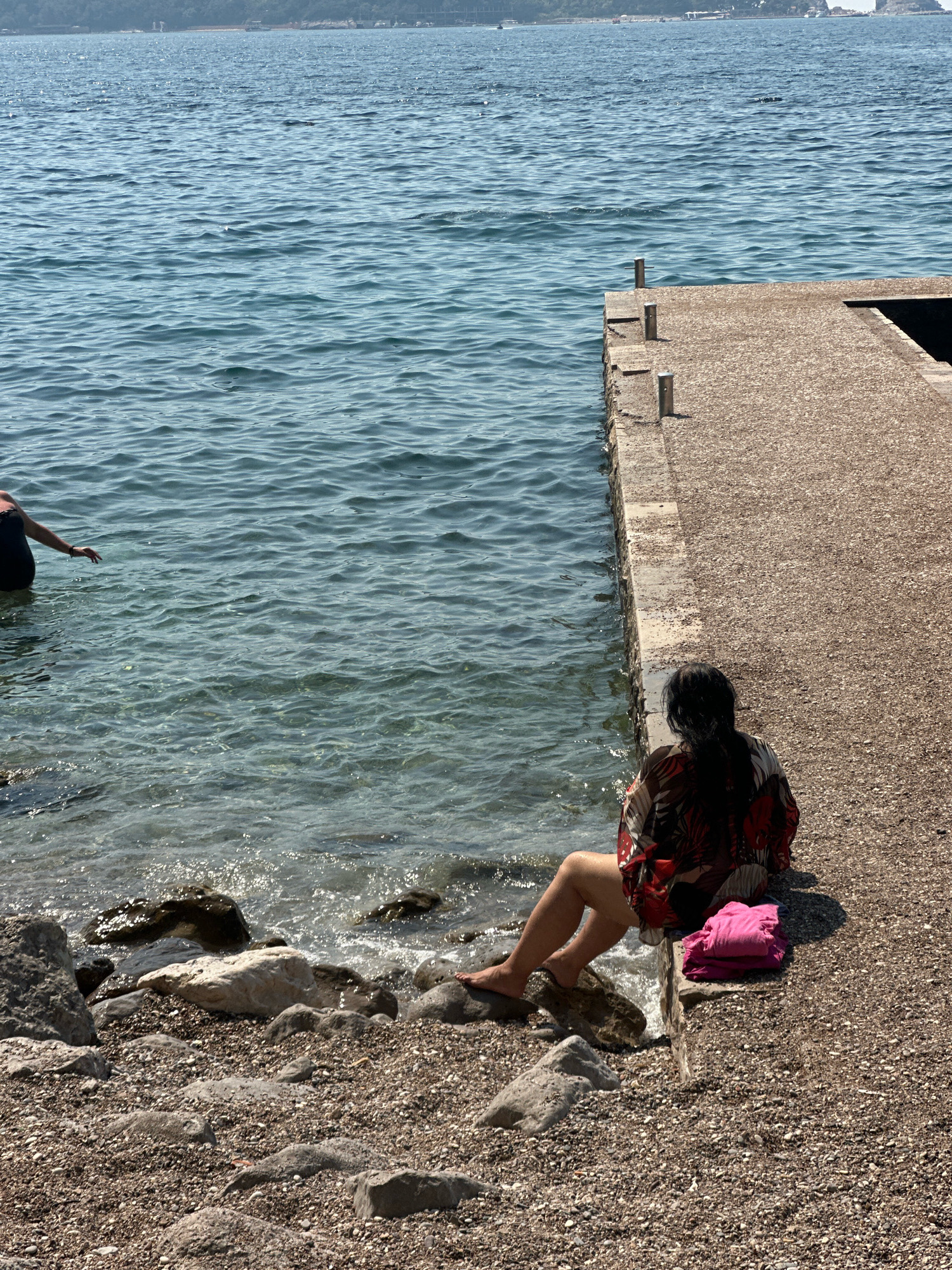 A woman with dark hair, wearing a patterned shirt and shorts, sits on a pebble beach next to a concrete pier. She is facing away from the camera and towards the sea. A pink towel is folded on the pier next to her. Another person, wearing a dark swimsuit, is partially visible in the water to the left. The sea is calm with gentle waves. In the background, a distant shoreline with buildings and trees can be seen. The scene appears to be in a coastal area of Budva, Montenegro.