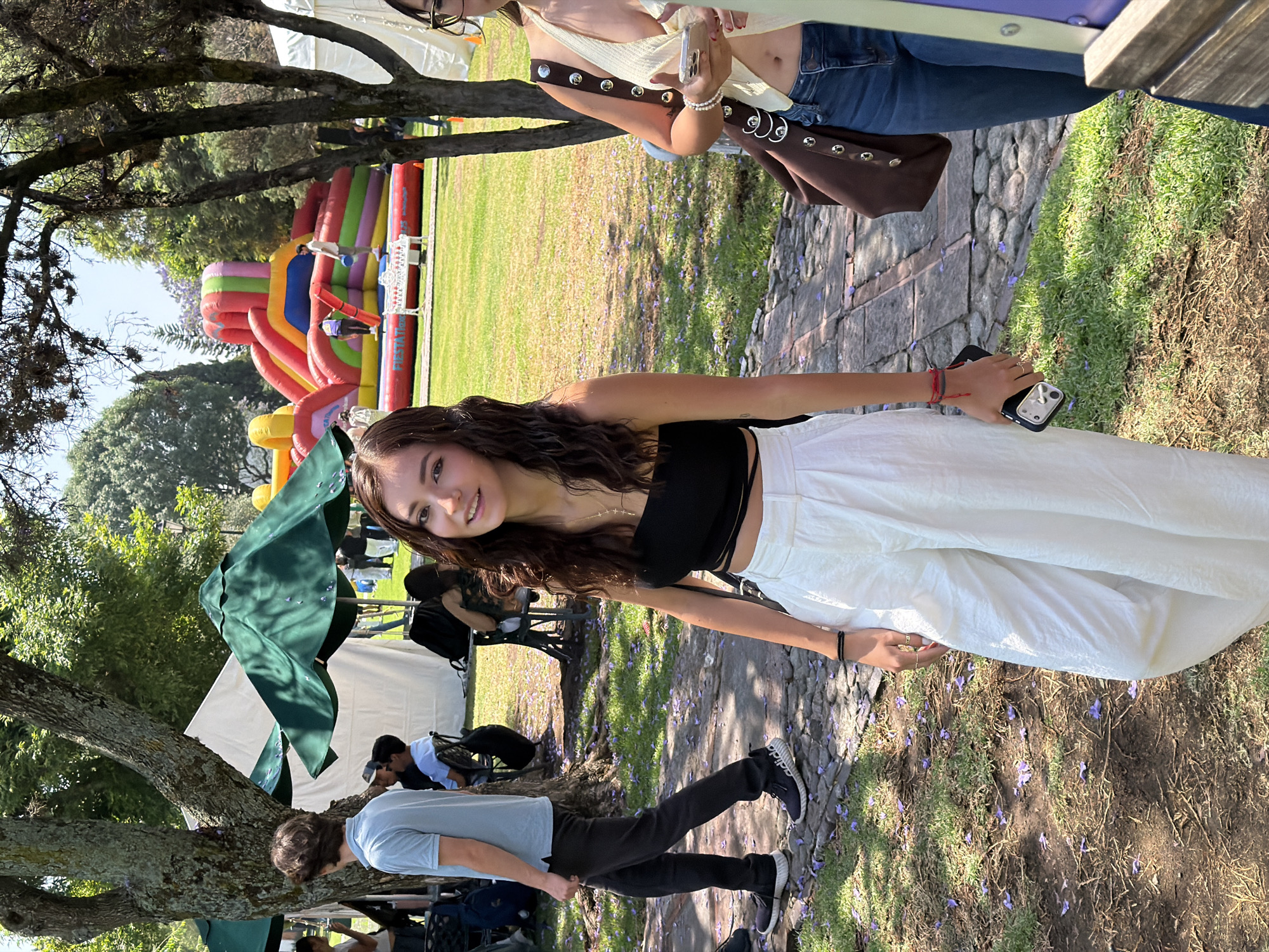 The image captures a vibrant outdoor gathering in San Andrés Cholula, Mexico, on a bright, sunny day. The central figure is a young woman with long, dark, curly hair, smiling directly at the camera. She is dressed in a black off-the-shoulder crop top and loose white wide-leg pants, paired with black sneakers. A red and black bracelet adorns her right wrist, and she holds a black smartphone with a silver camera unit in her hand.
In the background, a large, colorful inflatable play structure, possibly a bouncy castle or slide, dominates the grassy area with its red, yellow, blue, and green sections. Partially obscured by trees and tents, other people are visible, including a woman in a light top and jeans carrying a brown studded shoulder bag, and a man walking in a light blue t-shirt. Green tarpaulins and white tents suggest shaded areas for attendees.
The setting is a park-like environment with large trees providing dappled shade. The ground is a mix of green grass, dirt, and a stone-paved path, all liberally scattered with small purple petals, indicative of blooming Jacaranda trees common in the region. The overall atmosphere appears casual and festive. Visible text on the inflatable structure includes the word 'FIESTA'. Shadows indicate it is daytime, likely mid-morning or afternoon.