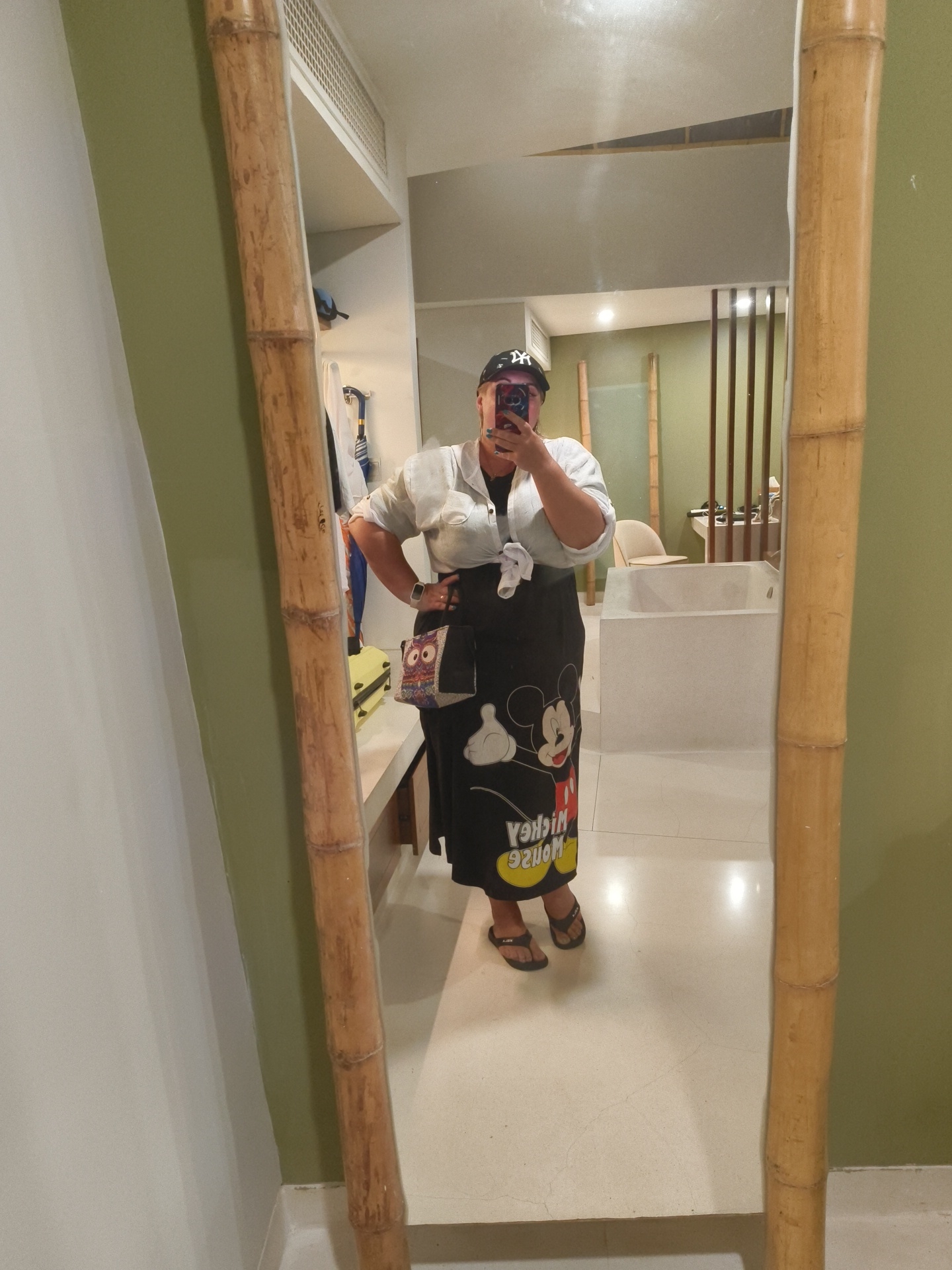 A woman, reflected in a large mirror with a bamboo frame, stands in what appears to be a hotel bathroom. She is wearing a white, loose-fitting shirt tied at the waist, a black baseball cap, and a long black skirt adorned with a large print of Mickey Mouse. She is holding a black smartphone in her right hand, taking the selfie. On her left hip, she has a small, square patterned bag. The bathroom features a large bathtub, a light-colored countertop, and a light-colored tiled floor. The walls are painted a pale green. The overall impression is that of a casual, possibly resort-style setting. The image does not contain any specific visual cues that would allow for definitive location identification within Phú Quốc, Vietnam, beyond the possibility of it being a hotel or resort.
