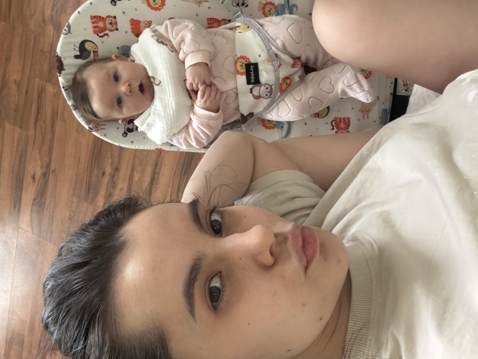 This is a high-angle, vertical shot capturing a woman and a baby indoors, likely in a home in Chaikovsky, Russia. The image is oriented such that the subjects appear rotated clockwise.
In the upper left, a baby lies in a patterned bouncer. The baby has fair skin, dark eyes, and light brown hair. They are wearing a light pink, long-sleeved onesie with heart patterns and a plain white bib. The baby's mouth is slightly open, and they look directly at the camera with an alert expression. Their hands are clasped together. The bouncer features various cartoon animals, including lions, tigers, owls, and toucans. A black label on the bouncer's restraint strap clearly reads "Baby&Joy" in white text.
The woman is positioned in the foreground, lying down with her head towards the bottom right of the frame. Her dark hair is pulled back, and she has dark eyebrows and eyes. She looks directly into the camera with a neutral expression. She is wearing a light-colored (possibly white or light beige) crew-neck top. Part of her bare arm or leg is visible near the baby in the bouncer.
The background consists of a wooden floor with prominent grain, visible around the bouncer and behind the woman's head. The lighting appears soft and natural, suggesting it is daytime indoors. The overall scene conveys a quiet, domestic moment.