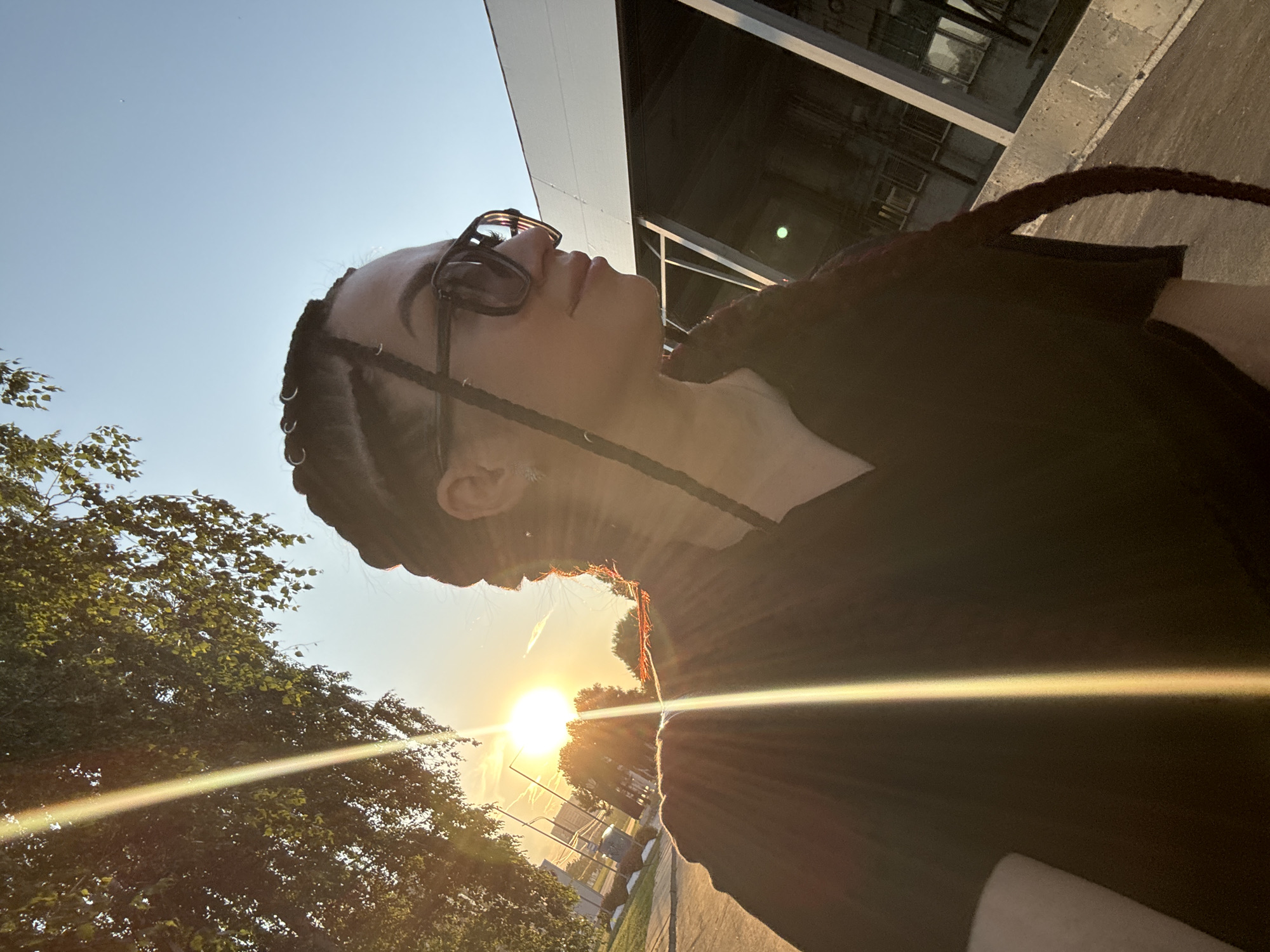 A young woman with braided hair and sunglasses is captured in a sun-drenched, outdoor setting. The sun is low in the sky, casting long rays of light across the scene. The woman is wearing a dark-colored v-neck top and is looking to the right of the frame. In the background, there's a building with a modern architectural design, featuring large glass panels and a sloped roof. Trees with green foliage are visible on the left side of the image, and the sky is a clear, pale blue. The ground appears to be paved, possibly a sidewalk or a road. The overall impression is that of a candid photograph taken during a sunset.