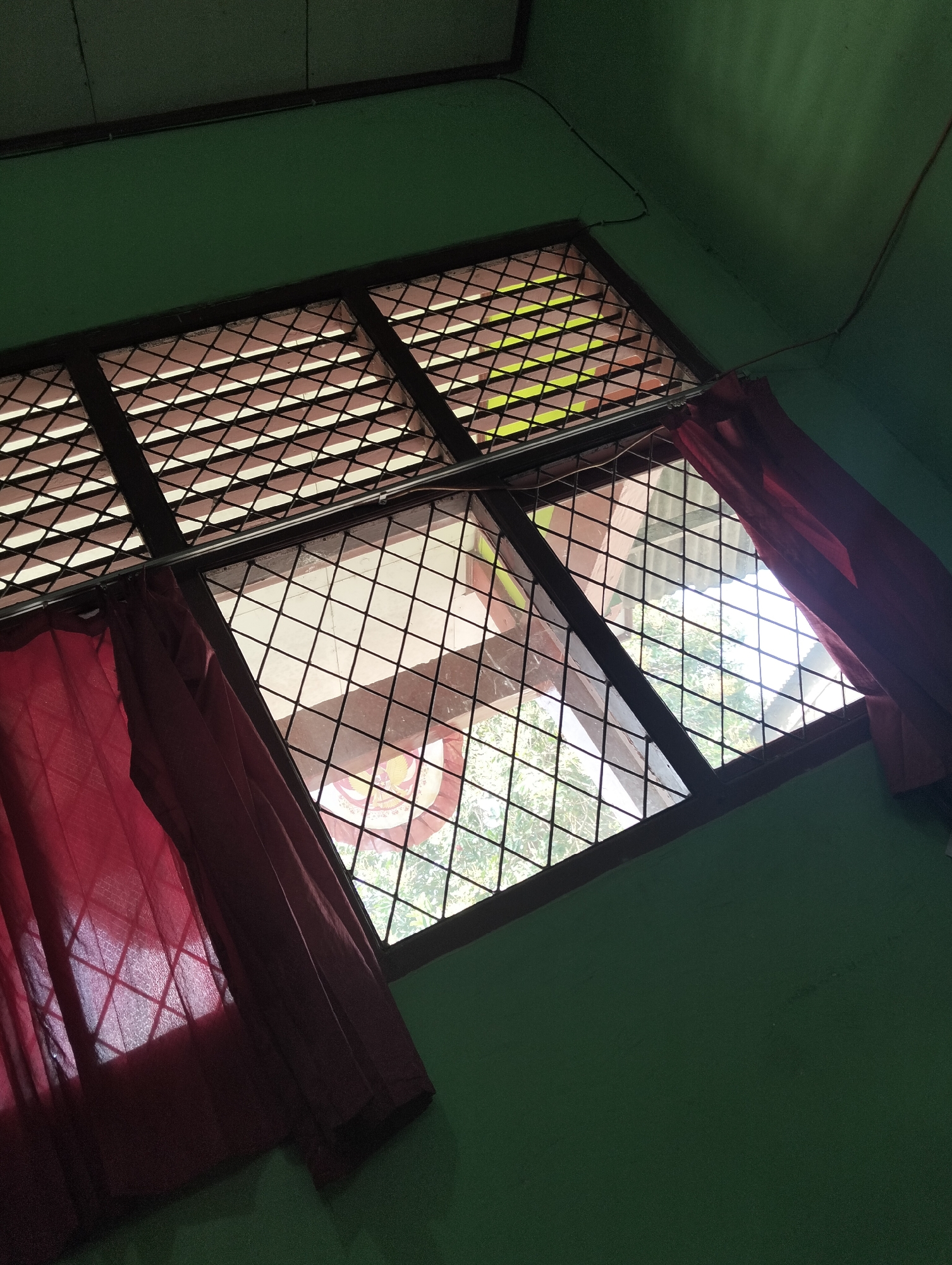 The image depicts an interior view of a room with a green painted wall and ceiling. A diamond-patterned window grate is visible, with red curtains partially drawn on the left and right sides. Light streams through the window, illuminating the grate and casting shadows. Outside the window, greenery and roofing structures are discernible. The scene appears to be a residential interior, potentially in Banjar Agung, Indonesia, characterized by its simple furnishings and natural light.