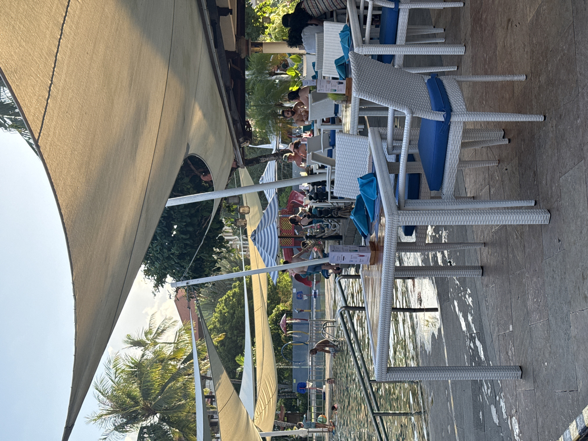 An outdoor dining area with white wicker tables and chairs with blue cushions is set up next to a swimming pool at a resort in Kuta Selatan, Indonesia. Several people are in the pool, some swimming and others relaxing. There are also people sitting at tables in the dining area, some eating and drinking. Palm trees and lush greenery surround the pool and dining area. The sky is clear and blue, and the sun is shining. A large, light-colored awning provides shade over the dining area. In the background, there are colorful water slides and playground equipment. The image captures a lively and relaxed atmosphere at a tropical resort.