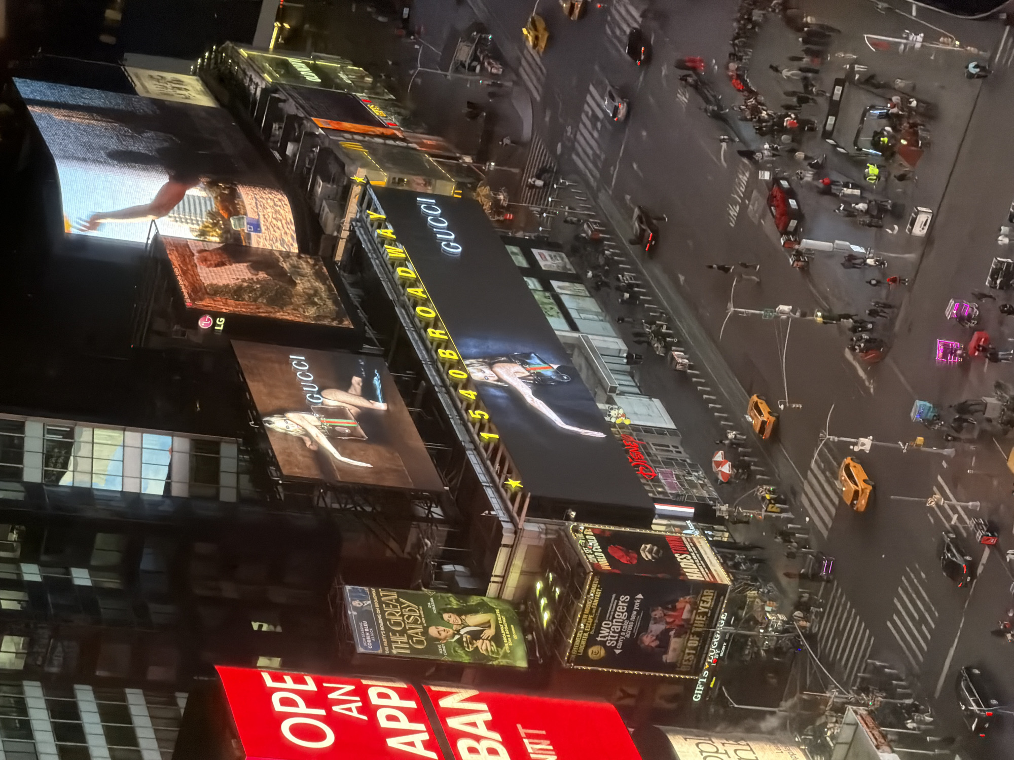 Here is a description of the image:
This is an aerial view of Times Square in New York City at night. The image captures the bustling street scene below, with a multitude of pedestrians visible on the sidewalks and crossing the streets. Yellow taxis are moving through the streets amidst the pedestrian traffic. High-rise buildings surround the square. There are numerous illuminated billboards. Prominent brands and shows like "Gucci," "The Great Gatsby," and advertisements from companies like "LG," and "Sony," are on the signs. The street is brightly lit from the signage and the buildings.