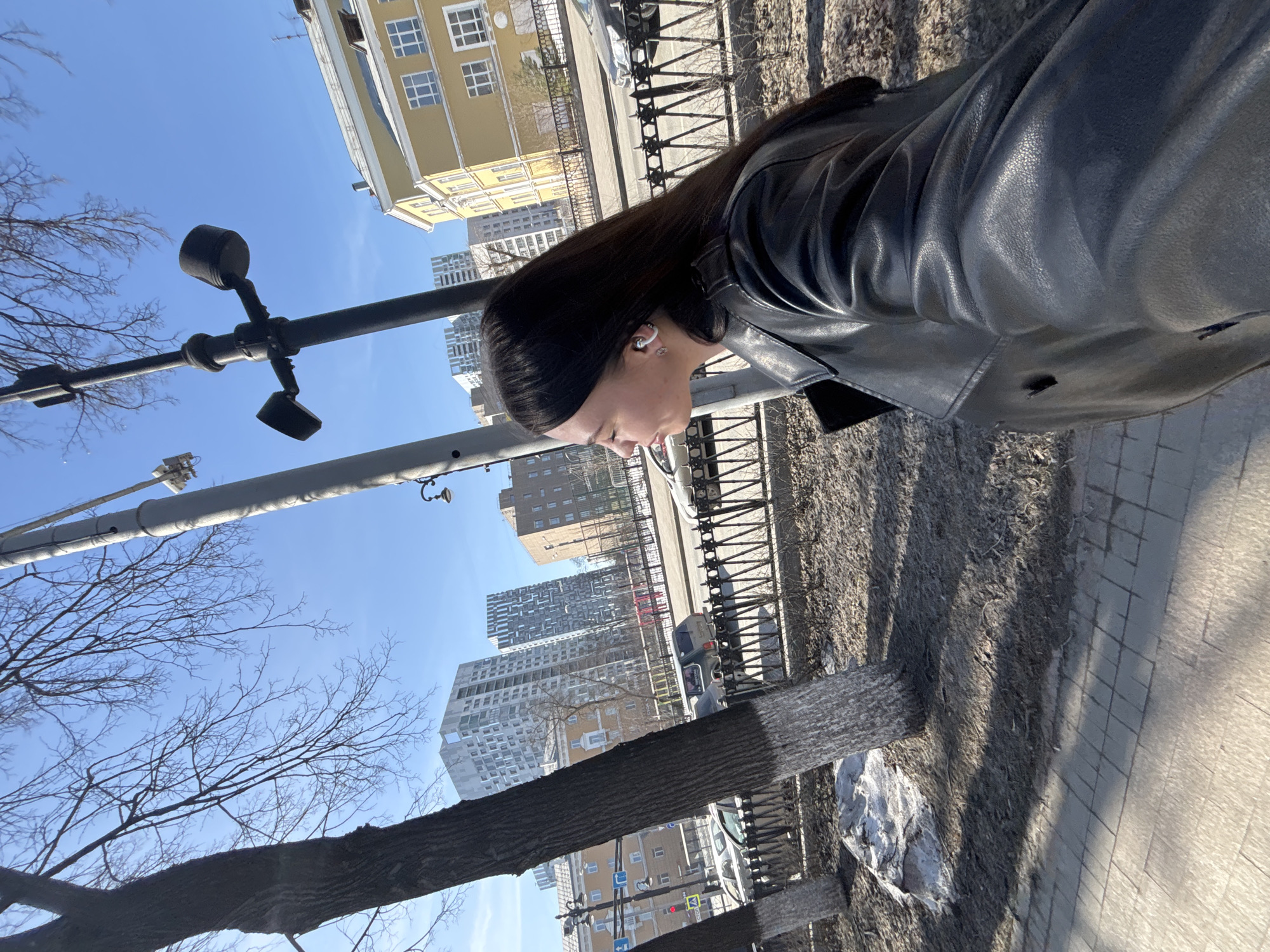 A woman with long dark hair and wearing a black leather jacket stands on a sidewalk in what appears to be Perm, Russia. Behind her, a black metal fence separates the sidewalk from the street, where cars are parked. The background features various buildings, including tall apartment complexes and an older yellow building with many windows. A tall black lamppost and bare tree branches are also visible. The sky is clear and blue, indicating a sunny day.