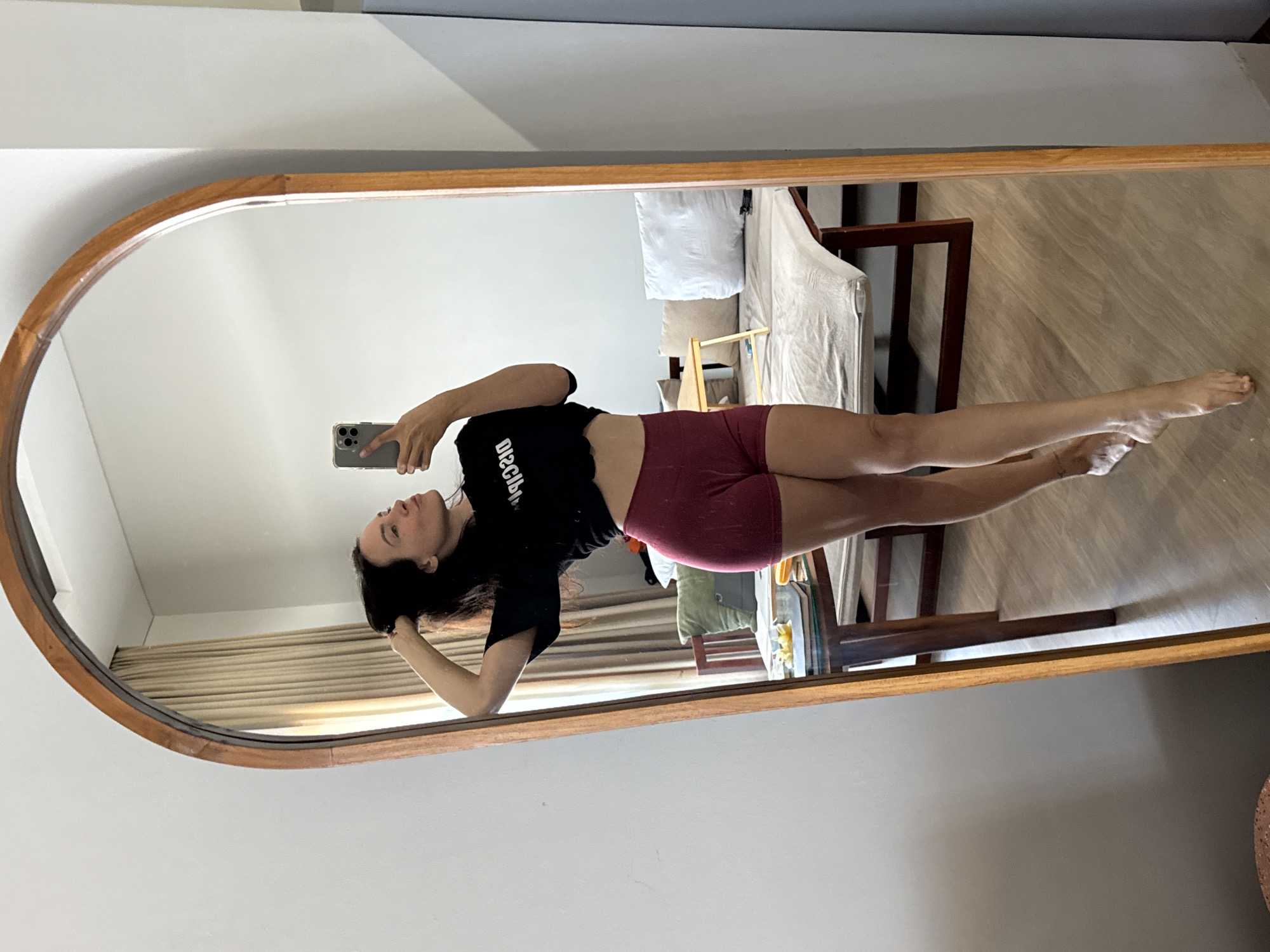 A woman is taking a selfie in a large, oval mirror. She is wearing a black crop top with white text and maroon shorts. She is standing with her left hand in her hair and her right hand holding her phone. The mirror reflects a room with a bed, a table, and curtains. The lighting suggests it is daytime. The location is North Kuta, Indonesia.