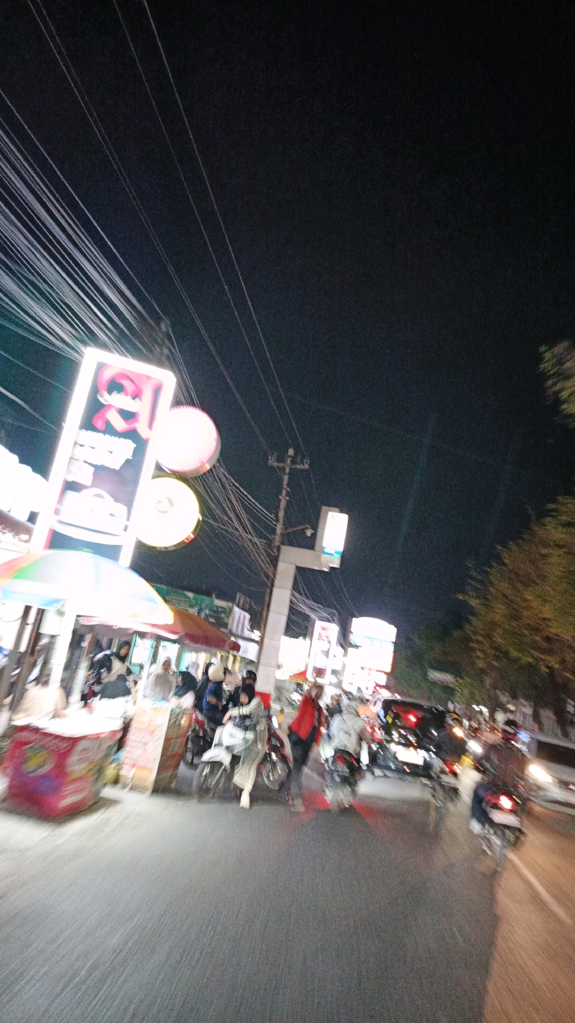 This is a nighttime photograph of a busy street scene in Pabelan, Indonesia. The image is characterized by motion blur, suggesting movement of both the camera and the subjects. The foreground is dominated by a blurry asphalt road. On the left side of the street, there are market stalls with umbrellas, and several people are visible, some appearing to be vendors and others customers.
Further down the street, there is significant traffic, primarily consisting of motorcycles with riders. Some of the motorcycles are moving, with their headlights and taillights creating streaks of light. Several individuals are riding on the motorcycles, many wearing traditional Islamic head coverings. A few cars are also present in the traffic.
On the left side of the street, there are tall, brightly lit signs. One prominent sign is rectangular and vertically oriented, displaying a pink logo with some text. To its right, there are two large, circular, illuminated objects, likely lanterns or part of a larger illuminated advertisement. In the background, there are utility poles with numerous electrical wires crisscrossing overhead. Further down the street, other illuminated signs are visible, contributing to the nighttime ambiance. The overall impression is a vibrant and active street scene in an Indonesian city at night.