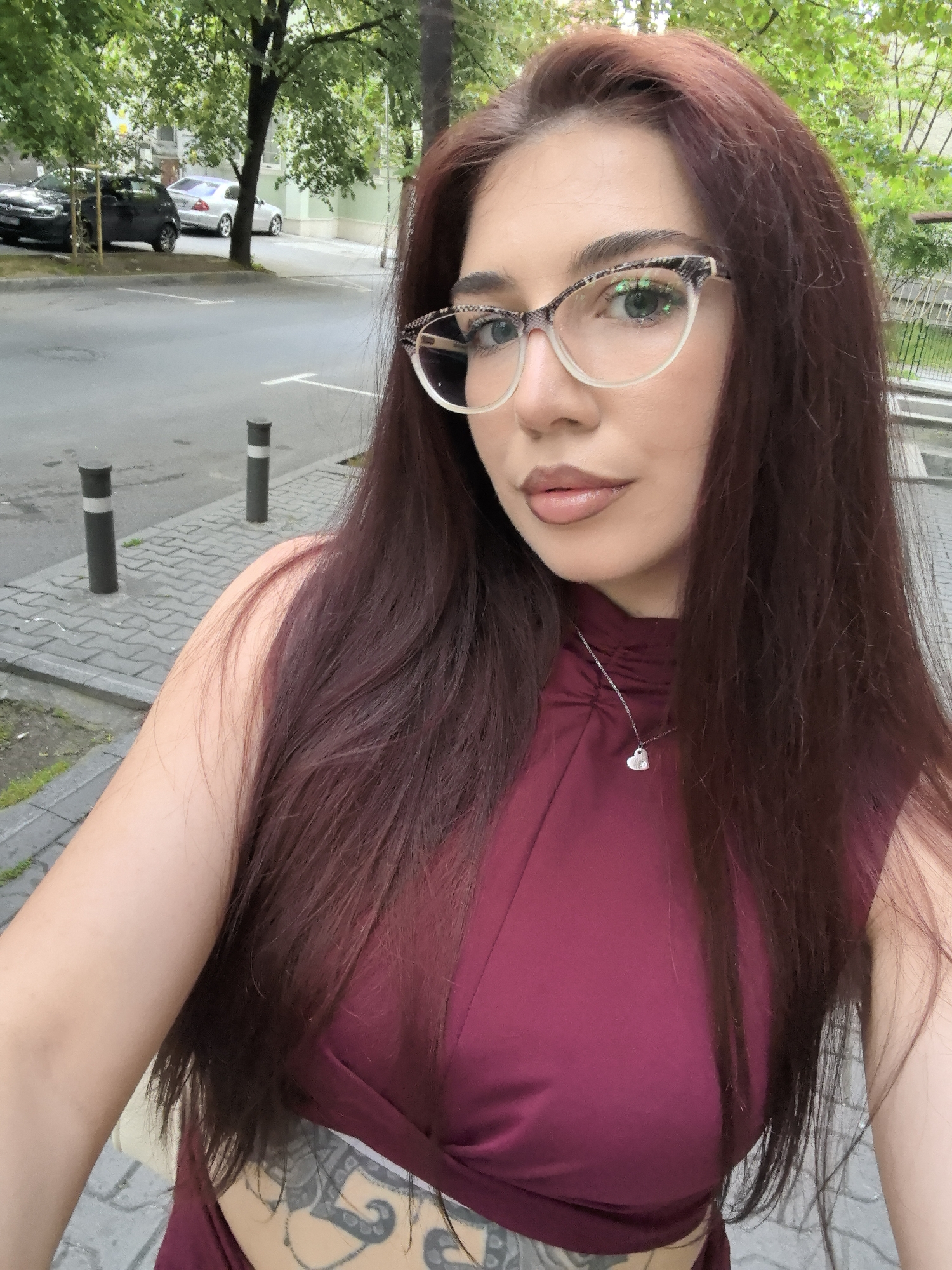 A woman is posing outdoors in Stara Zagora, Bulgaria. She has long, auburn hair and is wearing glasses with a patterned frame, a dark red dress, and a silver necklace with a heart pendant. Part of a tattoo is visible on her side.
Behind her, there's a street with parked cars and a sidewalk with bollards. Trees with green leaves are also in the background. The sidewalk is paved with interlocking gray concrete stones.