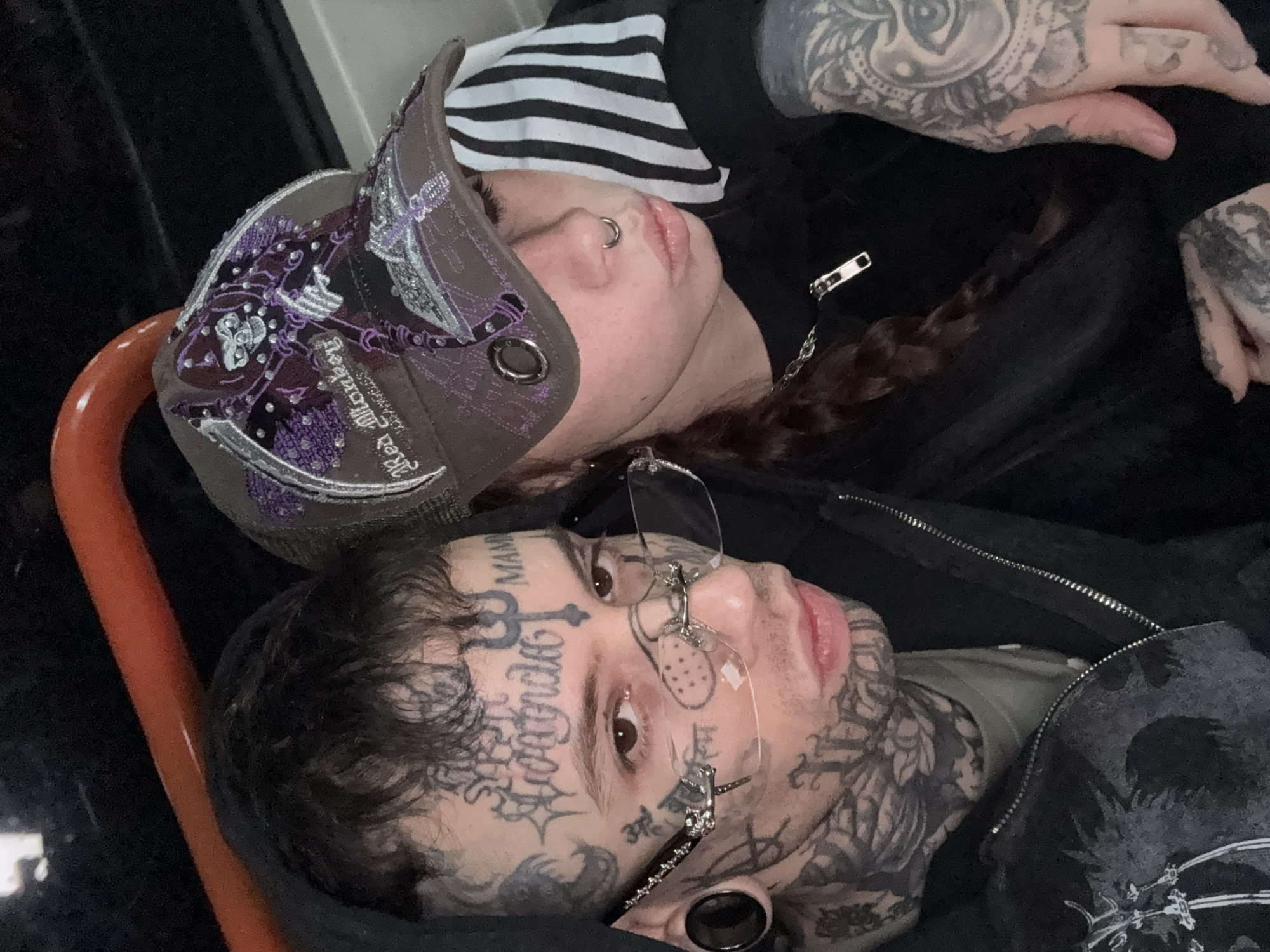 Within the interior of a public transport vehicle in Buenos Aires, Argentina, two heavily tattooed individuals are captured in a close-up, dim-light portrait.
The individual on the left, appearing male, has short dark hair and brown eyes. His face is extensively covered with black ink tattoos, including the word "Contreras" across the forehead, a trident symbol between the eyebrows, "MAMI" above the right eyebrow, and various other intricate designs around his eyes, cheeks, and jawline, extending onto his neck. He wears decorative rimless eyewear, featuring silver metal accents, with large black ear gauges in his earlobes. His lips are pursed, and he looks directly into the camera. He is dressed in a dark hooded jacket, partially unzipped, revealing a lighter garment underneath.
The individual on the right, appearing female, has long, dark reddish-brown hair styled in a braid. Her face is partially obscured by a brown trucker-style cap featuring elaborate purple and silver embroidery depicting a grim reaper, along with the text "Red Monkey Los Angeles." She has a silver hoop septum piercing. Her visible hands and fingers are heavily tattooed, including an eye motif on the back of her right hand. She wears a dark top with a silver chain necklace that has a zipper-pull pendant. A black and white striped garment is visible beneath or behind her. Her lips are slightly pursed, and her gaze is directed away from the camera.
An orange curved grab bar or handle is visible in the upper background, consistent with a vehicle interior. Both individuals are positioned closely, with the female leaning near the male, as they pose for the photograph. The specific location within Buenos Aires is not discernible from the image.