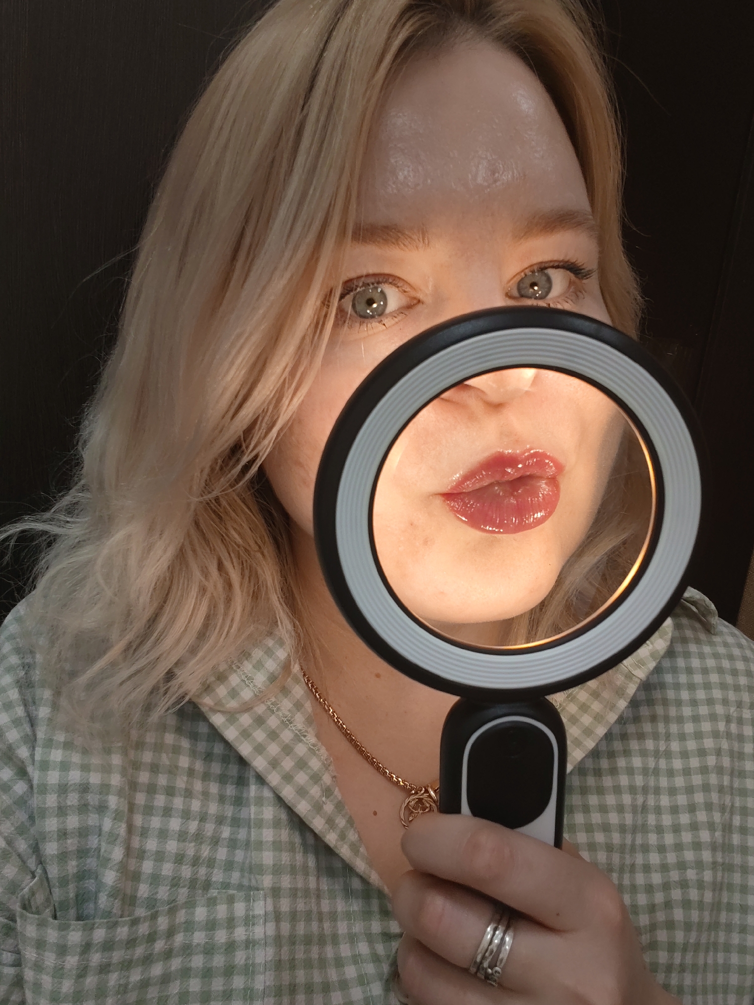 A woman with blonde wavy hair and blue eyes is holding a ring light up to her face, focusing the light on her glossy lips. She is wearing a green and white checkered shirt, and a gold chain necklace. Her hand, adorned with three rings, is holding the ring light. The background is dark, possibly a room with dark furniture. There is no information available to place this scene in Izhevsk, Russia.