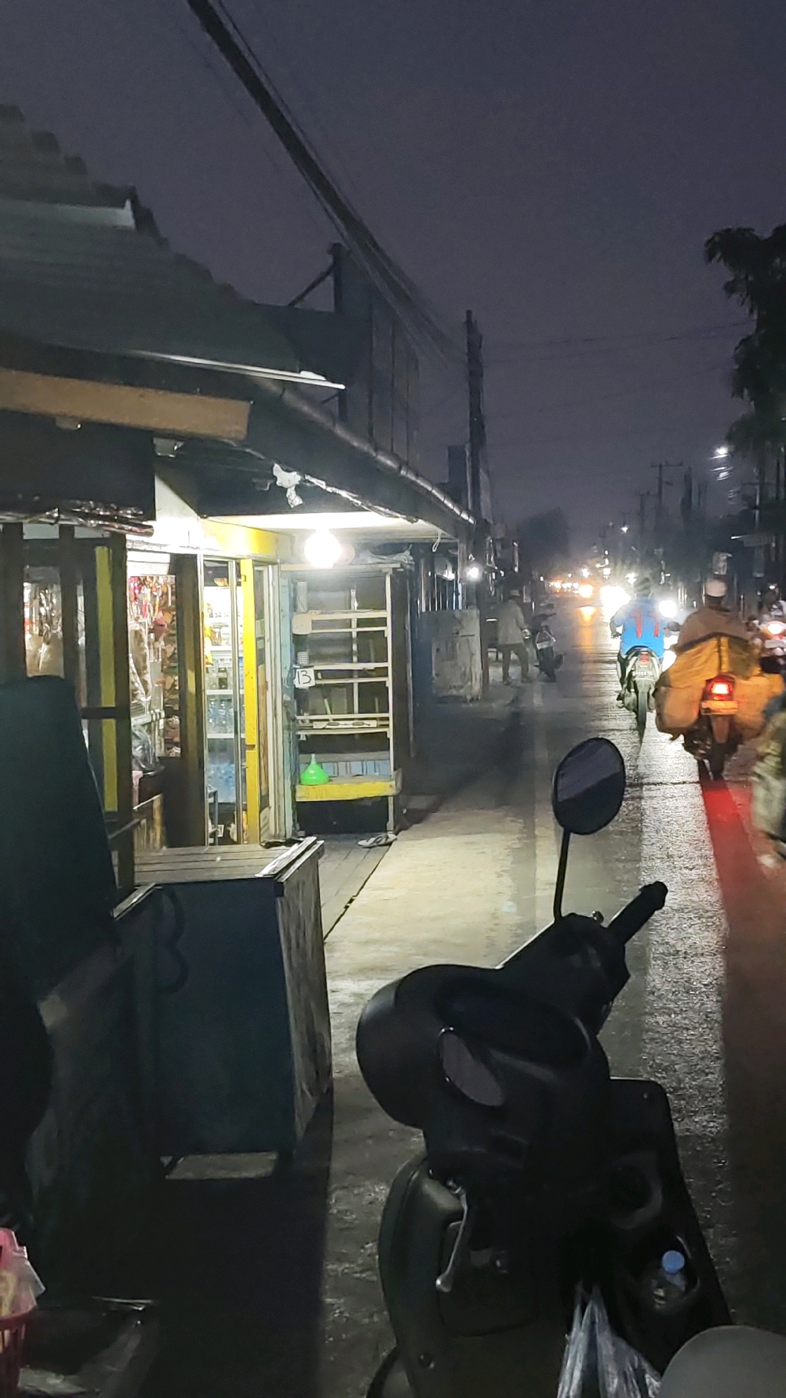 At night in Sungai Lulut, Indonesia, a street scene unfolds with dimly lit shops and a wet asphalt road reflecting streetlights. In the foreground, a black scooter is parked, facing away from the camera. The scooter's handlebars and front fairing are visible, with a side mirror attached to the left handlebar. A water bottle cap is visible in a compartment on the scooter. To the left, a dark, rectangular object, possibly a bin or a stall, is positioned along the sidewalk. Further down the street, several motorcycles are traveling, some carrying passengers and cargo, indicated by the loads strapped to the back. The shop on the left side of the street is illuminated by warm interior lighting, showcasing shelves stocked with various items behind glass doors. One shelf in an open display unit contains a green plastic container. The road appears wet, suggesting recent rain, and the sky is dark and overcast. Streetlights and the headlights of passing vehicles illuminate the scene.