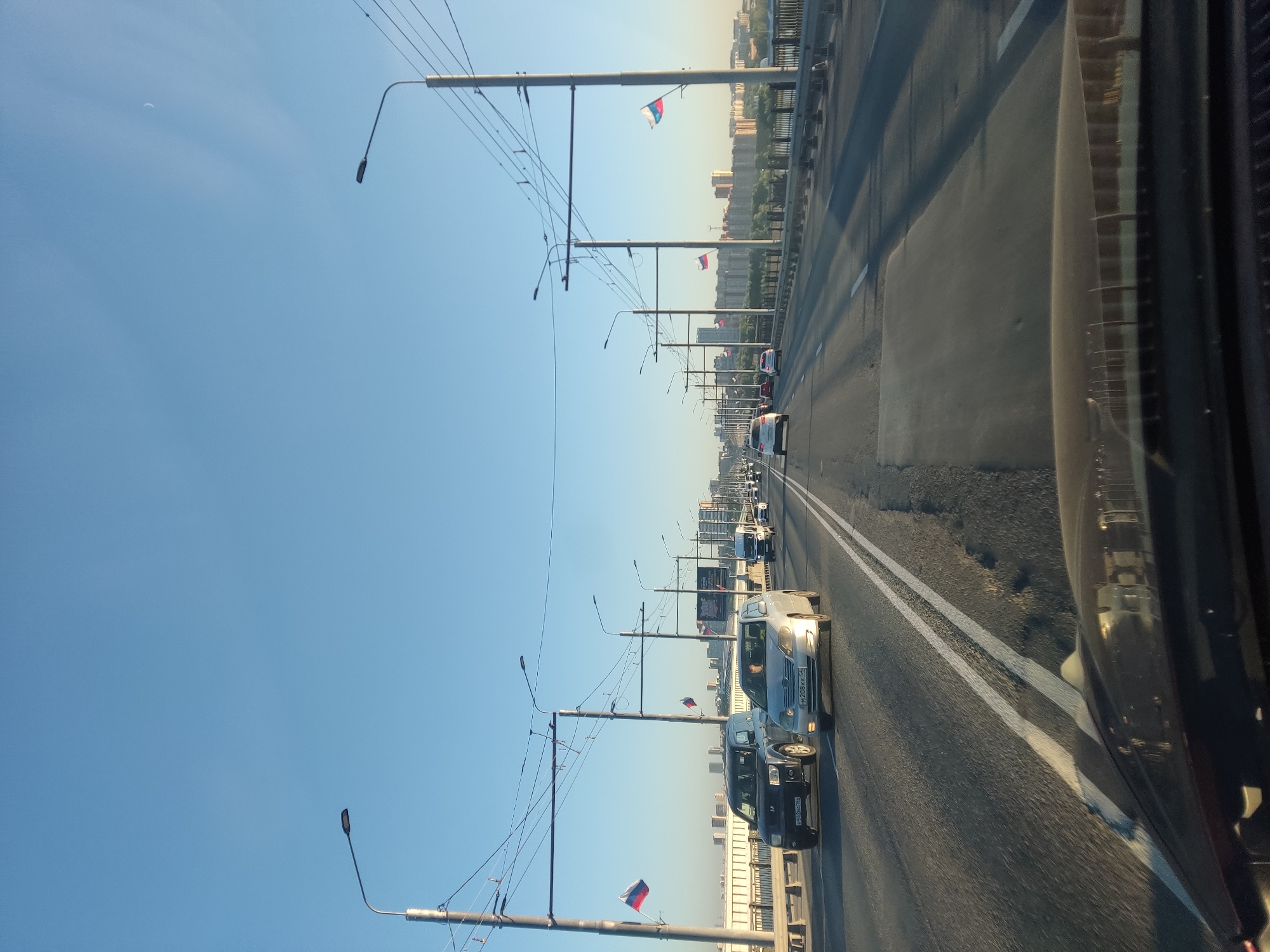 The image shows a view from a moving vehicle driving across a bridge, likely the Bugrinsky Bridge, in Novosibirsk, Russia. The sky is clear blue with a faint crescent moon visible. The bridge has multiple lanes of traffic, with several cars and a van visible. Russian flags are mounted on poles along the bridge. In the distance, buildings and a cityscape can be seen. The road surface appears worn in places. The time of day appears to be morning or late afternoon due to the lighting.