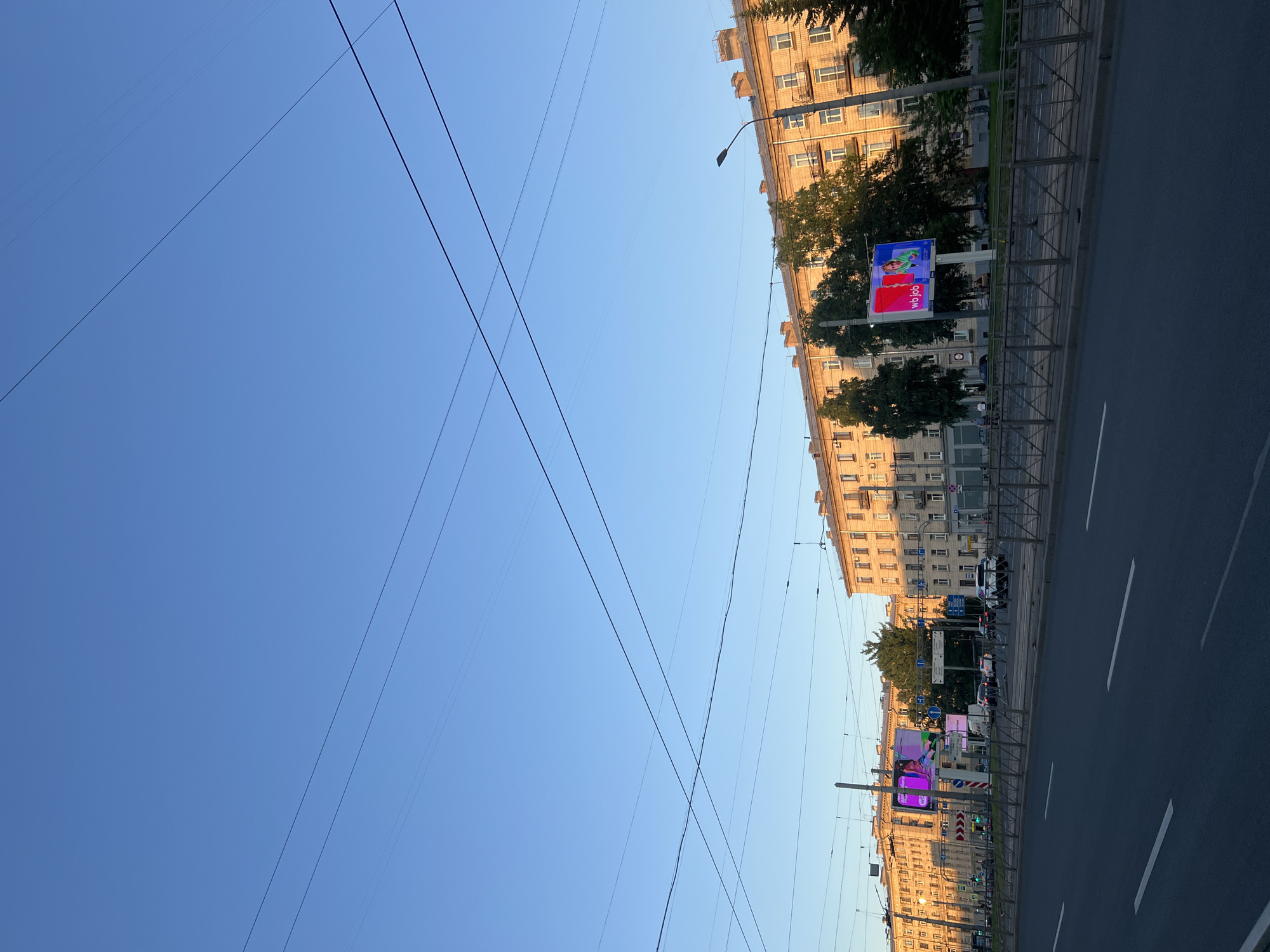This image depicts a wide urban streetscape in Saint Petersburg, Russia, specifically along Komendantsky Prospekt, likely near its intersection with Prospekt Ispytateley, looking generally west-southwest. The upper two-thirds of the frame is dominated by a clear, bright blue sky, traversed by numerous thin, dark overhead wires, consistent with public transport infrastructure.
Below the sky, multi-story residential buildings with light-colored, likely pale yellow or beige facades, line both sides of the wide avenue. The buildings on the right receive direct, warm, low-angle sunlight, creating a golden illumination and distinct shadows, while those on the far left are less directly lit. These buildings feature multiple windows and some balconies.
The foreground presents a multi-lane asphalt road with clear white dashed lane markings. A central median strip separates the traffic directions. On this median, two large digital billboards are prominently displayed. The billboard on the left shows abstract, brightly colored graphics, while the one on the right clearly displays "wb job" in white text on a colorful red and blue/green background, featuring a stylized human figure.
Various vehicles, including cars and a white SUV, are visible on the road, some in motion and others parked. A white delivery van is also present near the billboards. Several small, green trees are planted along the median strip and in front of the buildings. Street furniture includes dark streetlights, pedestrian railings, and multiple street signs. One blue directional sign, partially visible, indicates "НОВОЕ" (Novoye - New) and "КОМЕНД." (Komendantsky), referring to the Komendantsky Aerodrom district. A red traffic light with green elements is visible on the far left. A few pedestrians are discernible on the sidewalks or near the median, primarily on the left side of the road.