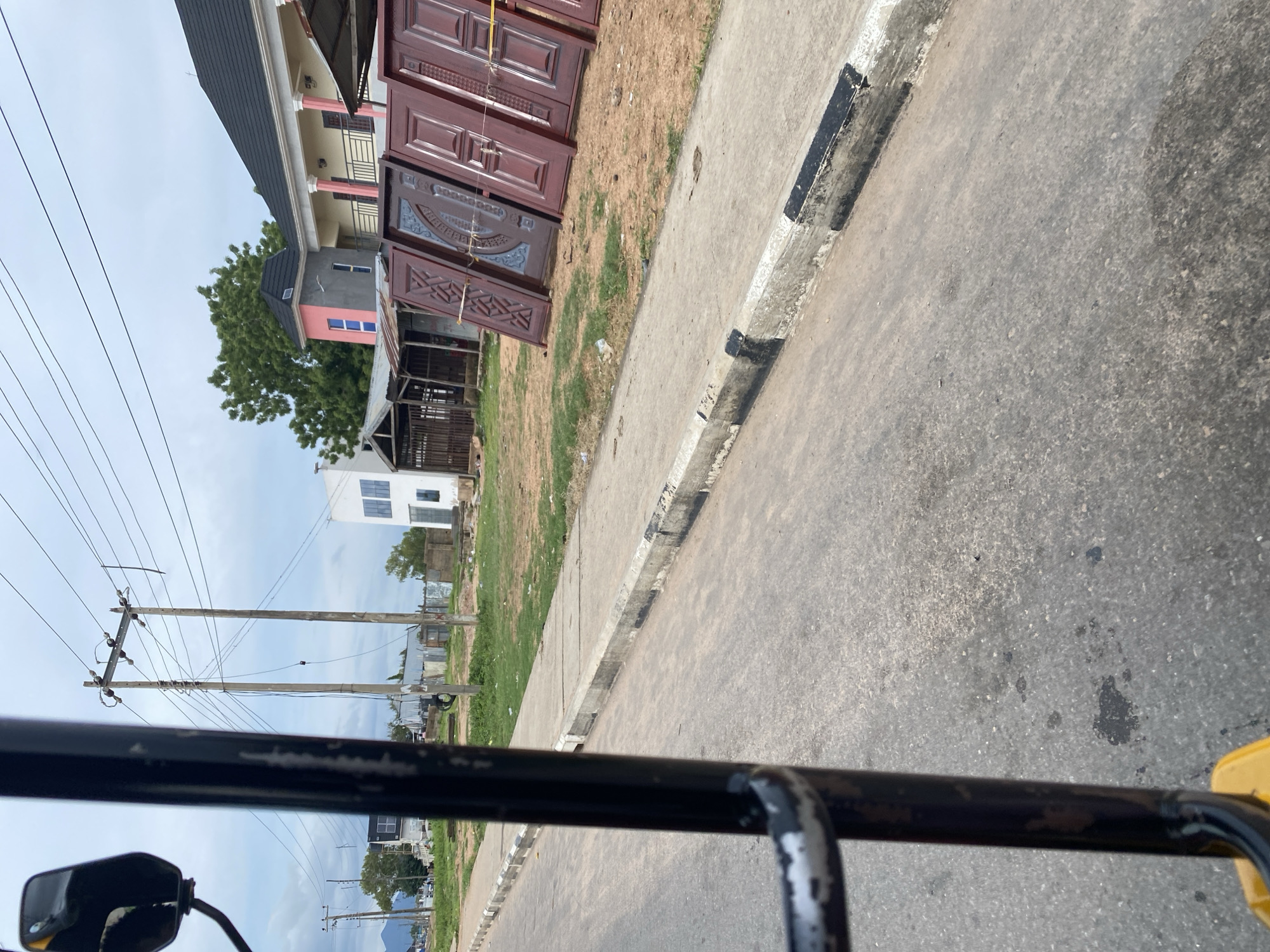 The image, taken from the perspective of a vehicle, captures a street scene in Jalingo, Nigeria. The foreground features the textured asphalt of a road, bordered by a concrete sidewalk with black and white painted edges. Beyond the sidewalk, a grassy area with scattered dirt and debris leads to a row of buildings. To the right, several large, ornate doors, likely for sale, are propped up against a structure. Behind them, a multi-story building with a light yellow facade, white columns, and a pink accent on its upper section is visible. Further back, a simpler white building with blue-tinted windows stands. Electrical poles with wires crisscross the sky, suggesting an active power infrastructure. The overall environment appears to be a commercial or residential area undergoing development or with open-air sales.