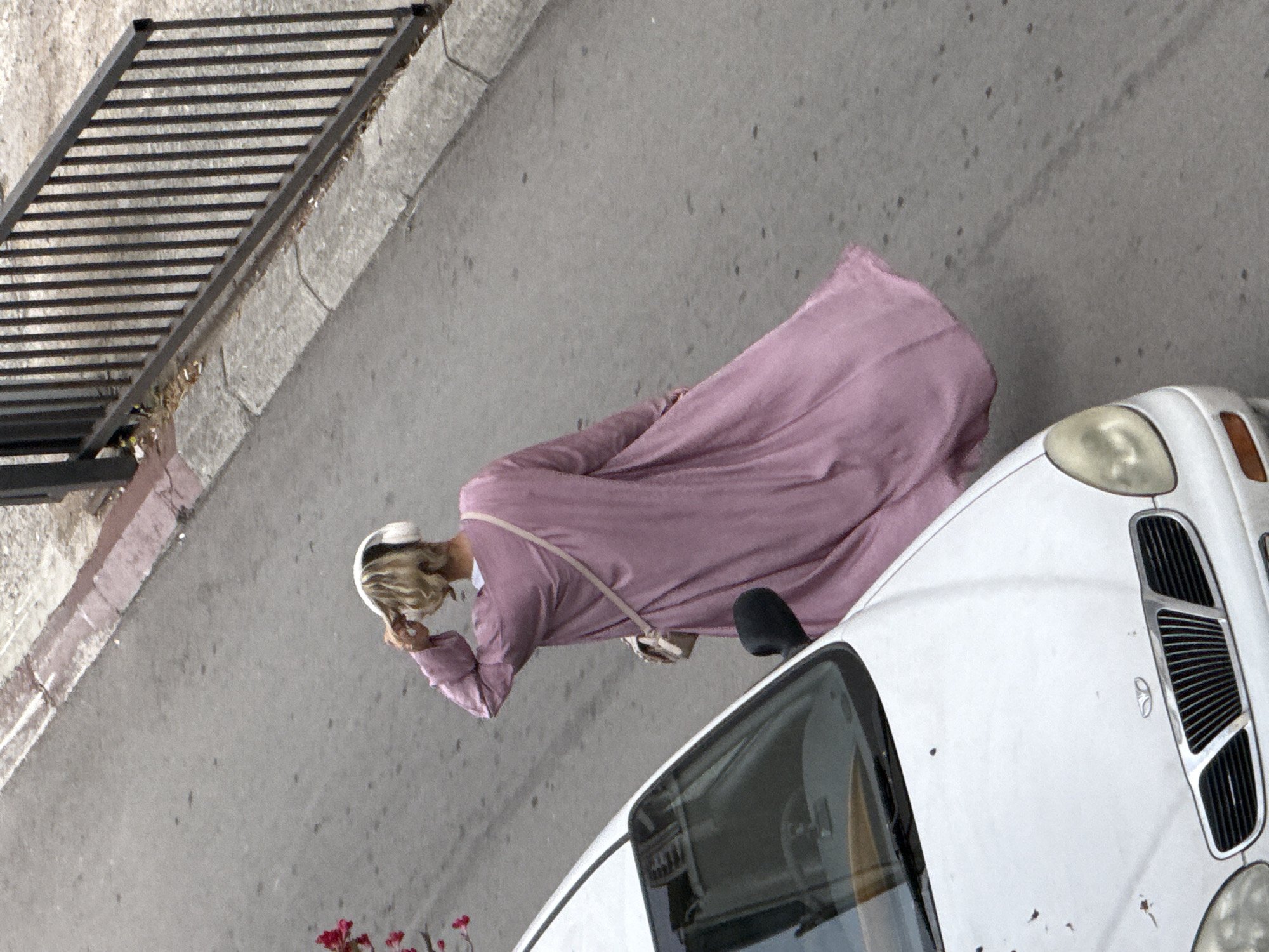 A woman with blonde hair is walking away from the camera on a street in Budva, Montenegro. She is wearing a long, pink, flowing dress and white headphones. She has a small purse slung across her body. A white car is parked to her left. A metal fence and curb are visible in the background.