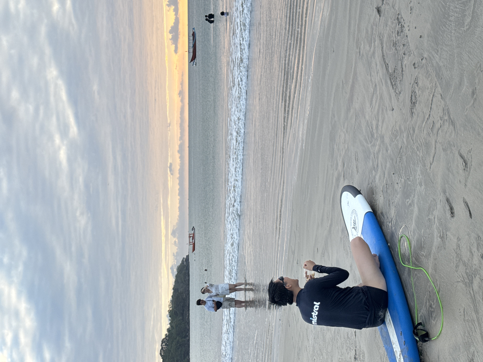An overcast sky with a gradient of pale blue to soft orange hues suggests either dawn or dusk at a beach in Kuta Selatan, Indonesia. The foreground features a person sitting on a blue surfboard on the sand, facing away from the camera and looking towards the ocean. This individual is wearing a black long-sleeved rash guard with the white logo "mistral" visible and black swim trunks. They are holding a clear cup in their right hand. A green surfboard leash is coiled on the sand beside the surfboard.
In the shallow water of the ocean, a couple stands looking at their phones, partially submerged. Further out, several other people are visible in the water. A red and blue boat with a white structure on top is anchored in the middle distance. The water is calm with gentle waves lapping at the shore. The beach appears to be a mix of sand and some damp, rippled sand where the water has receded. In the background, a tree-covered hillside forms the coastline.