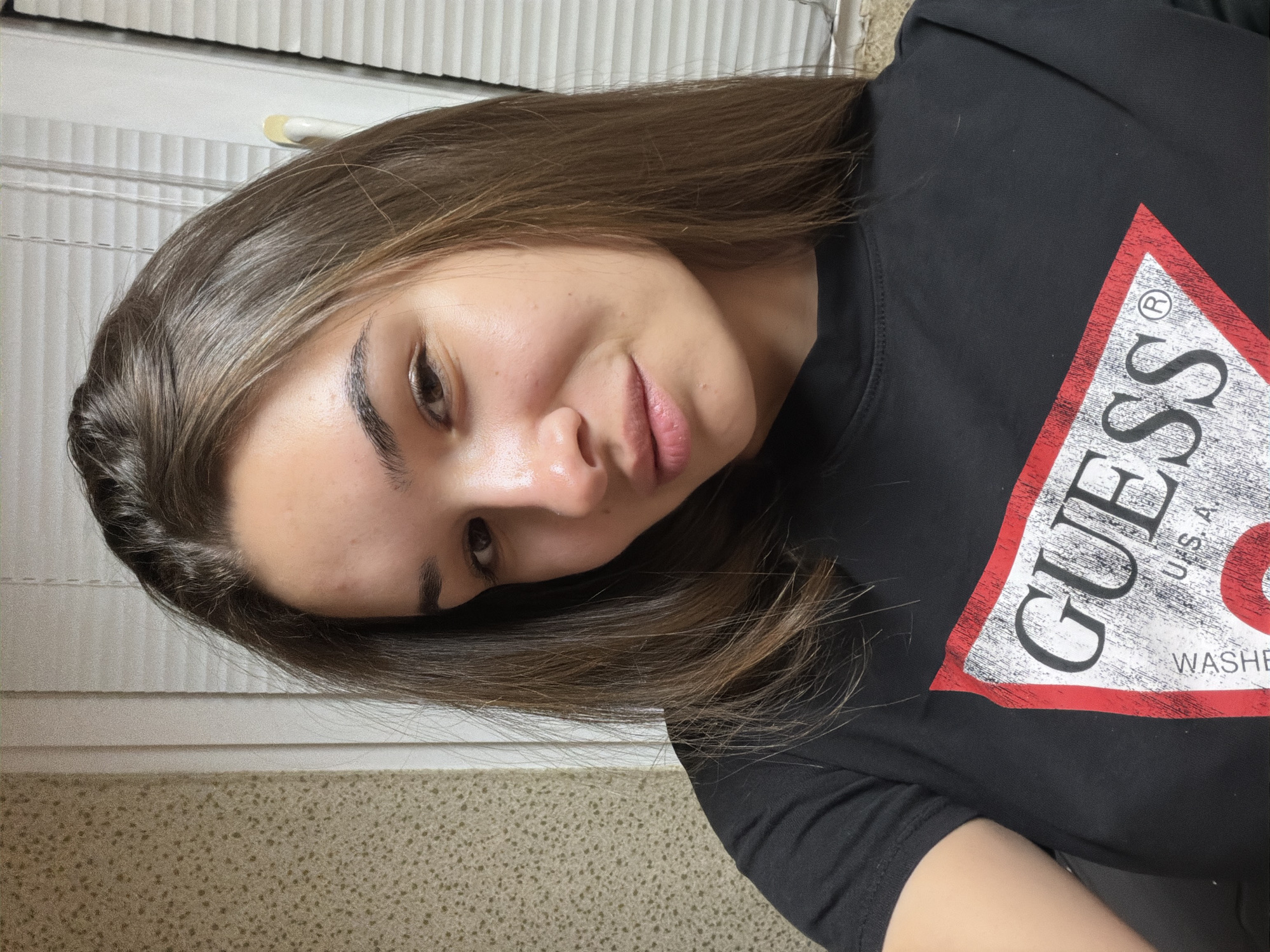 A person with brown hair is wearing a black t-shirt with a large "GUESS" logo. The person is looking at the camera with a slight smile. The background includes a textured wall and a white, corrugated surface, possibly a door or window covering. The location in Sofia, Bulgaria, is not discernible from the image.