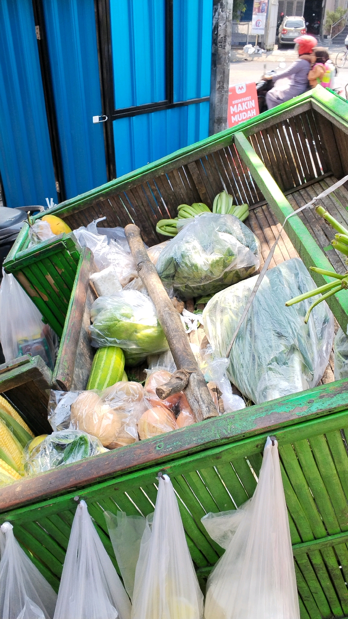 In the city of Banjaragung, Indonesia, a wooden cart filled with fresh vegetables and fruits is parked in front of a blue corrugated metal structure. The cart, painted green, is overflowing with produce, some of which is bagged in clear plastic. Among the visible items are bundles of leafy greens, cucumbers, corn on the cob, and what appear to be gourds or pumpkins. A wooden pole is propped up in the center of the cart, seemingly acting as a support. In the background, a street scene unfolds with a few cars and a motorcycle carrying two people, one of whom is wearing a red helmet. A sign advertising a delivery service is also visible on a pole. The overall impression is of a vibrant market scene on a bright day.