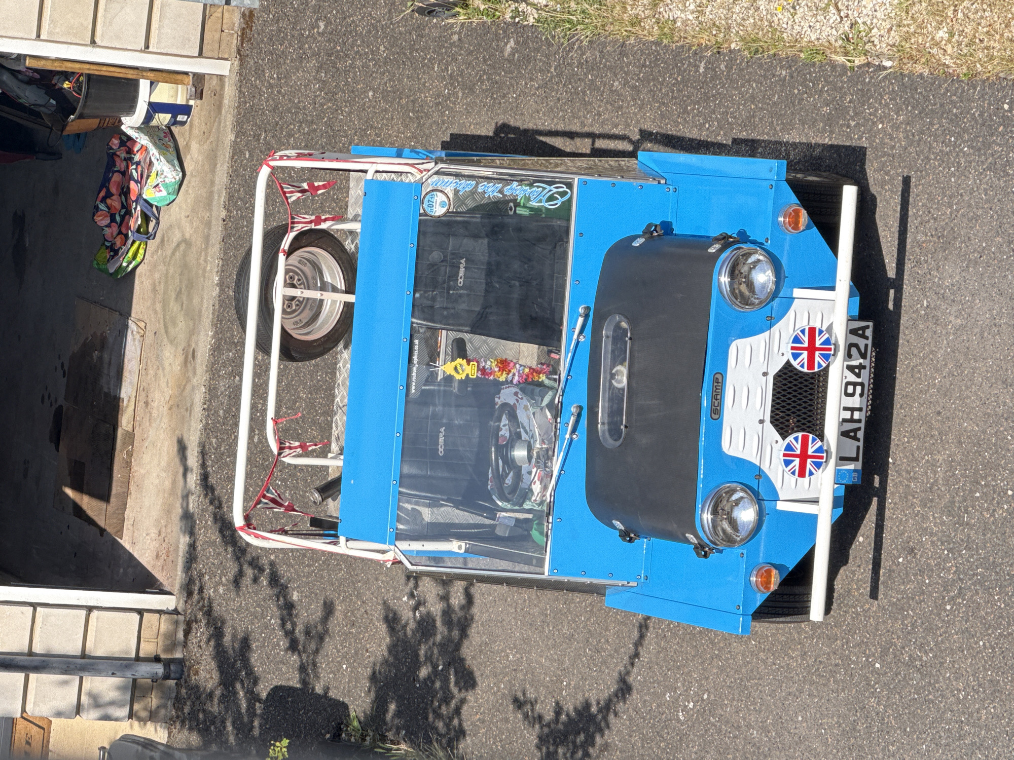 The image shows a bright blue Moke car parked on an asphalt driveway in Great Yarmouth, United Kingdom. The car has a white roll cage and a spare tire mounted on the rear. The front of the car features two round headlights, a grille with a Union Jack emblem, and a license plate that reads "LAH 942A". The car also has a black bonnet and windshield.
The driveway appears to be attached to a garage, with a concrete floor and a light-colored brick wall visible on the left. The scene is bathed in bright sunlight, casting shadows from the car and overhead trees. In the background, a black bucket and some colorful bags are visible near the garage entrance. The overall impression is of a sunny day in a residential area.