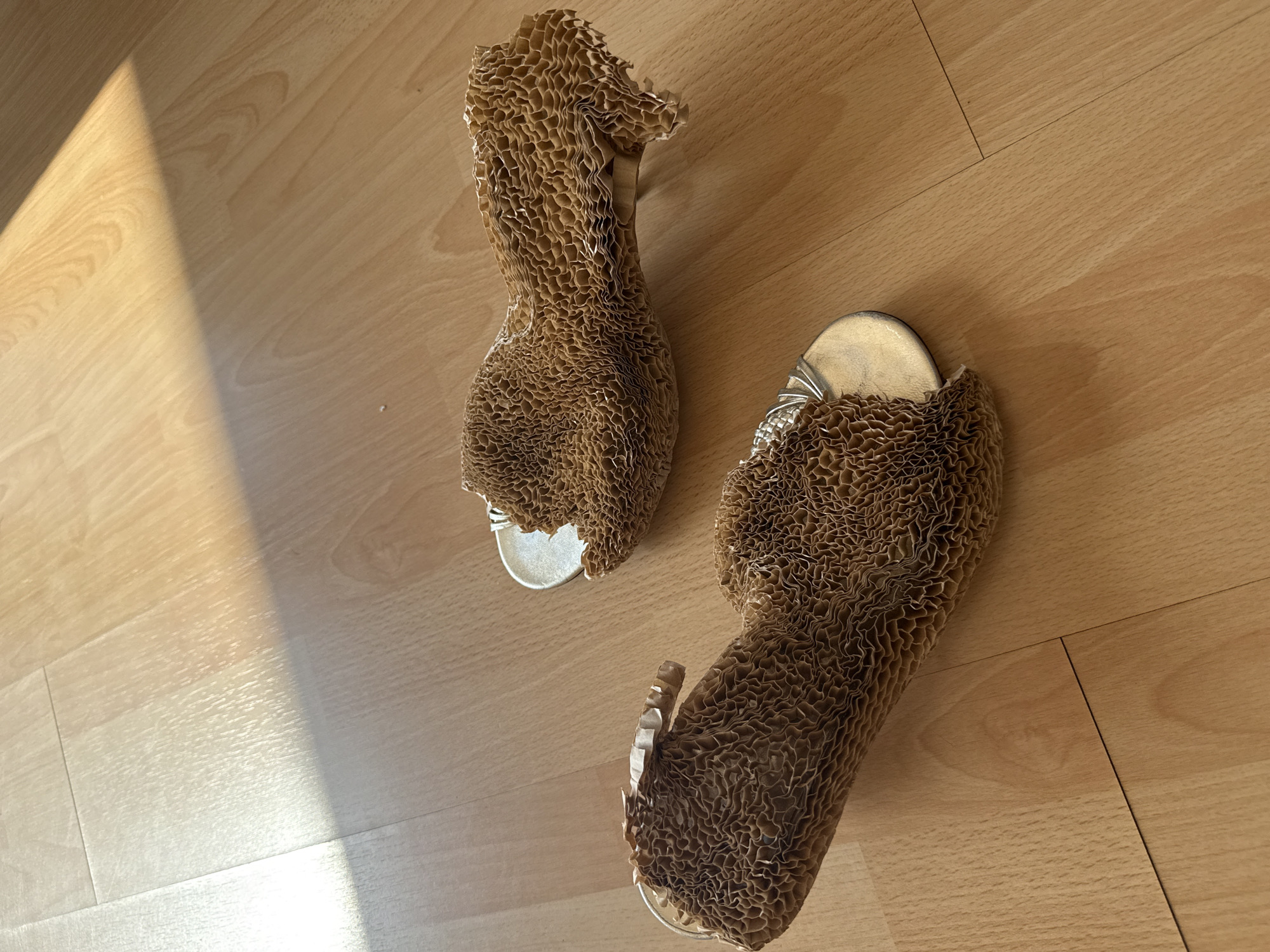 The image shows two high-heeled shoes on a light-colored wood floor. The shoes have a metallic silver toe and heel, and are covered in brown, cellular cardboard. The shoes are placed at an angle to one another, one pointing upwards and one pointing to the left. The floorboards run horizontally. A beam of sunlight falls on the left side of the image. The location is not identifiable as the city of London, United Kingdom, from the information in the image.