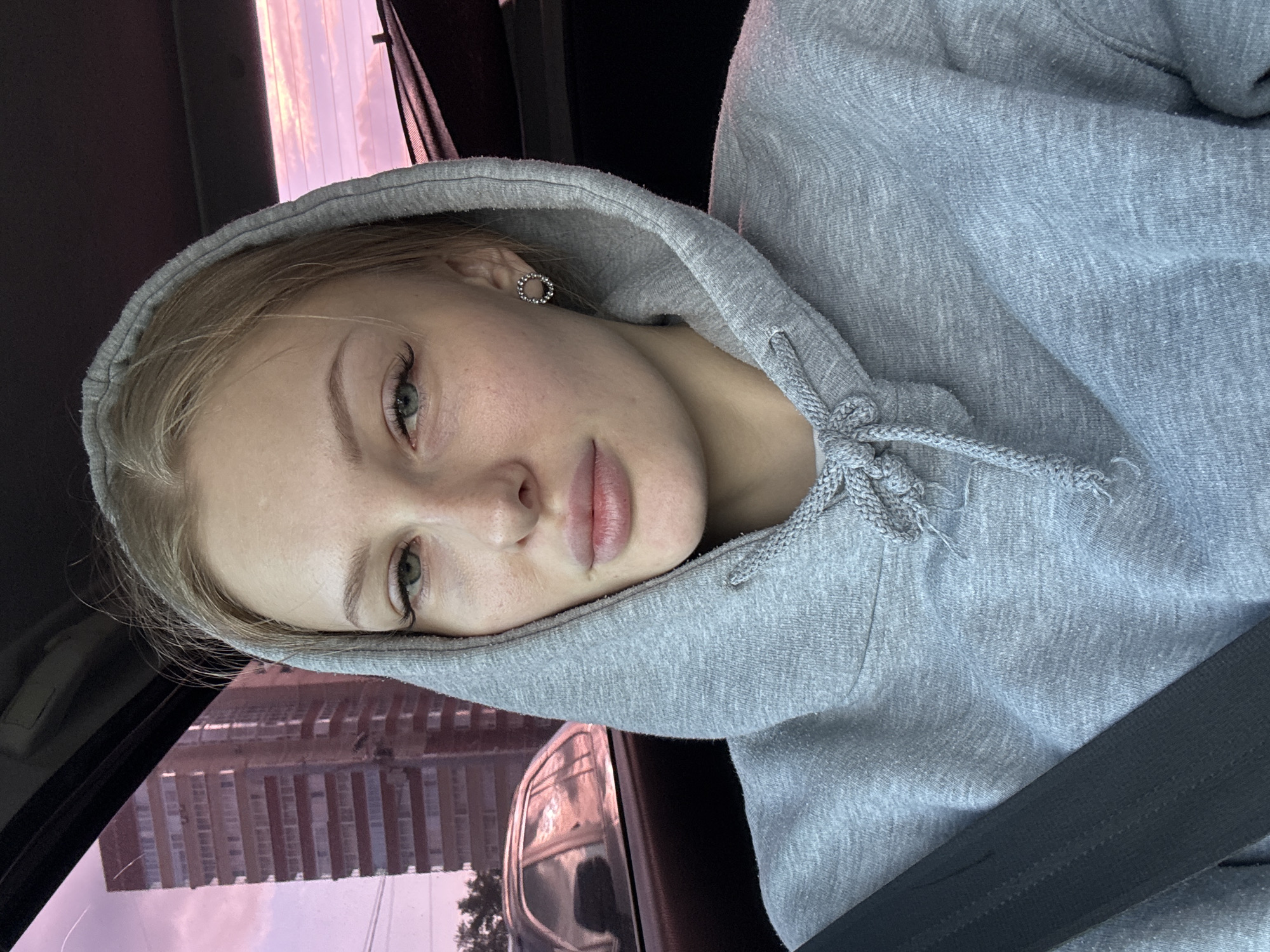 This media file is a close-up, rotated selfie of a young woman wearing a grey hooded sweatshirt. She is inside a vehicle, with a seatbelt visible across her chest. The setting appears to be urban, with a building and a cloudy, pink-hued sky visible through the car window. The time of day seems to be around sunset or sunrise due to the colors of the sky. The woman has blonde hair, light-colored eyes, and is wearing a small, circular earring. Her expression is neutral, and she is looking towards the camera. The image is well-lit, suggesting it was taken during daylight hours. There is no visible text in the media file.