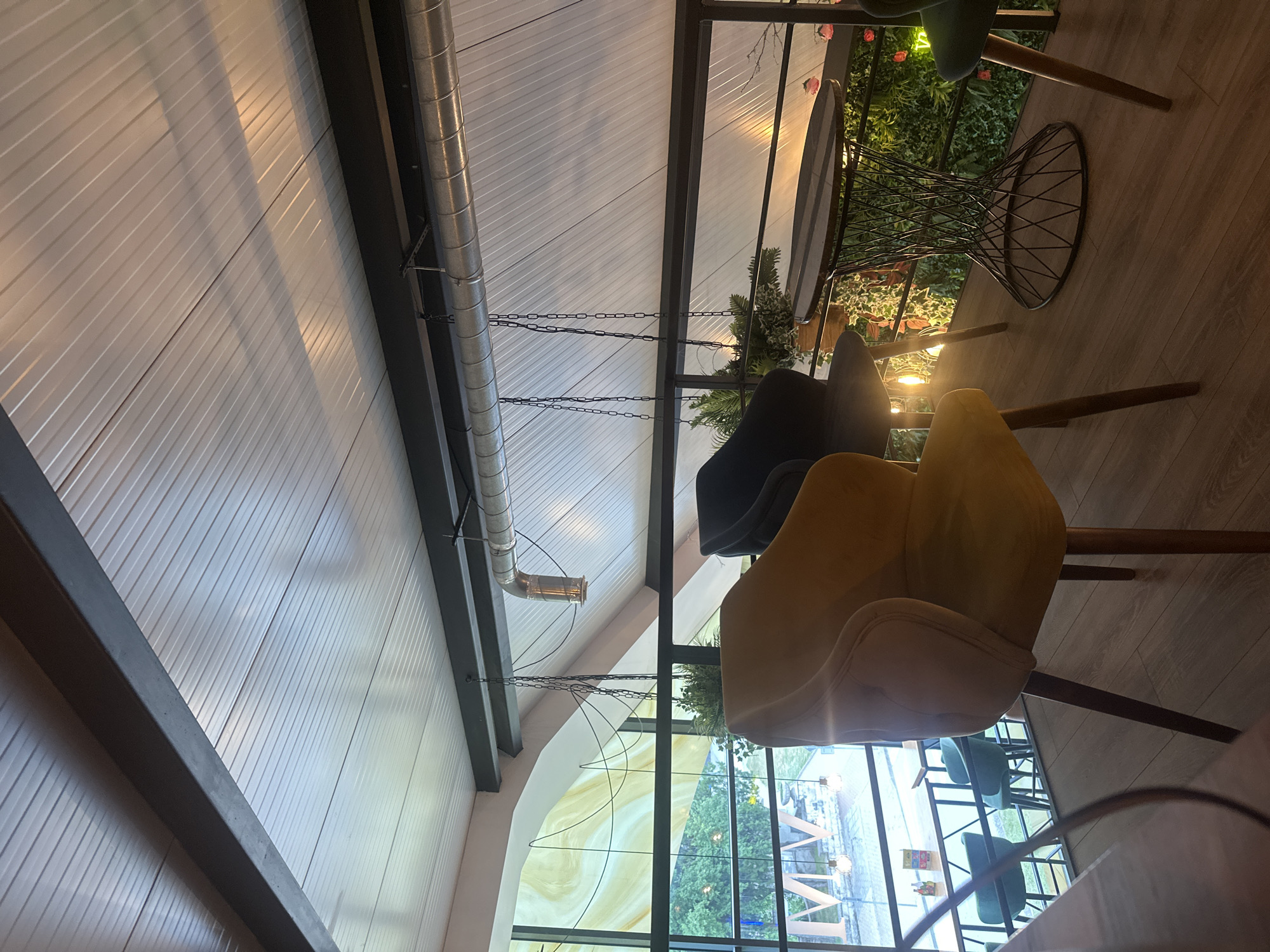 The image depicts the interior of a modern cafe, likely located in the city of Shumen, Bulgaria. The perspective is from a slightly elevated position, looking down and across a seating area.
The foreground features two comfortable upholstered chairs. The chair on the left is a vibrant yellow, with a plush, textured fabric and wooden legs. The chair on the right is a dark charcoal gray, also with wooden legs. Between these chairs and slightly to the right is a small, round coffee table with a dark, patterned top and a distinctive base made of thin, twisted black metal. Behind this table, a wall covered in artificial greenery with red flowers is visible, creating a biophilic design element.
In the mid-ground, a large window or glass partition dominates the background, offering a view of the exterior. Through the window, trees and streetlights can be seen, suggesting an urban setting. The window itself has a decorative, marbled effect in yellow and cream hues. Several smaller, teal-colored chairs are visible through the window, arranged at tables, suggesting additional seating within the cafe.
The ceiling is a prominent feature, constructed of white, ribbed panels, and supported by dark metal beams. A large, silver ventilation pipe runs horizontally across the ceiling, with a spotlight hanging from it. Two dark metal chains are suspended from the ceiling, one with a small potted plant attached.
The flooring is made of light-colored wooden planks. The overall ambiance is contemporary and cozy, with warm lighting creating a welcoming atmosphere. The scene is devoid of any visible people, suggesting a quiet moment in the cafe. The presence of plants, both real and artificial, adds a natural touch to the man-made environment. The architectural style suggests a renovated or purpose-built space designed for a casual dining or social experience. The exterior view through the window indicates it is likely daytime or dusk, given the lighting conditions.