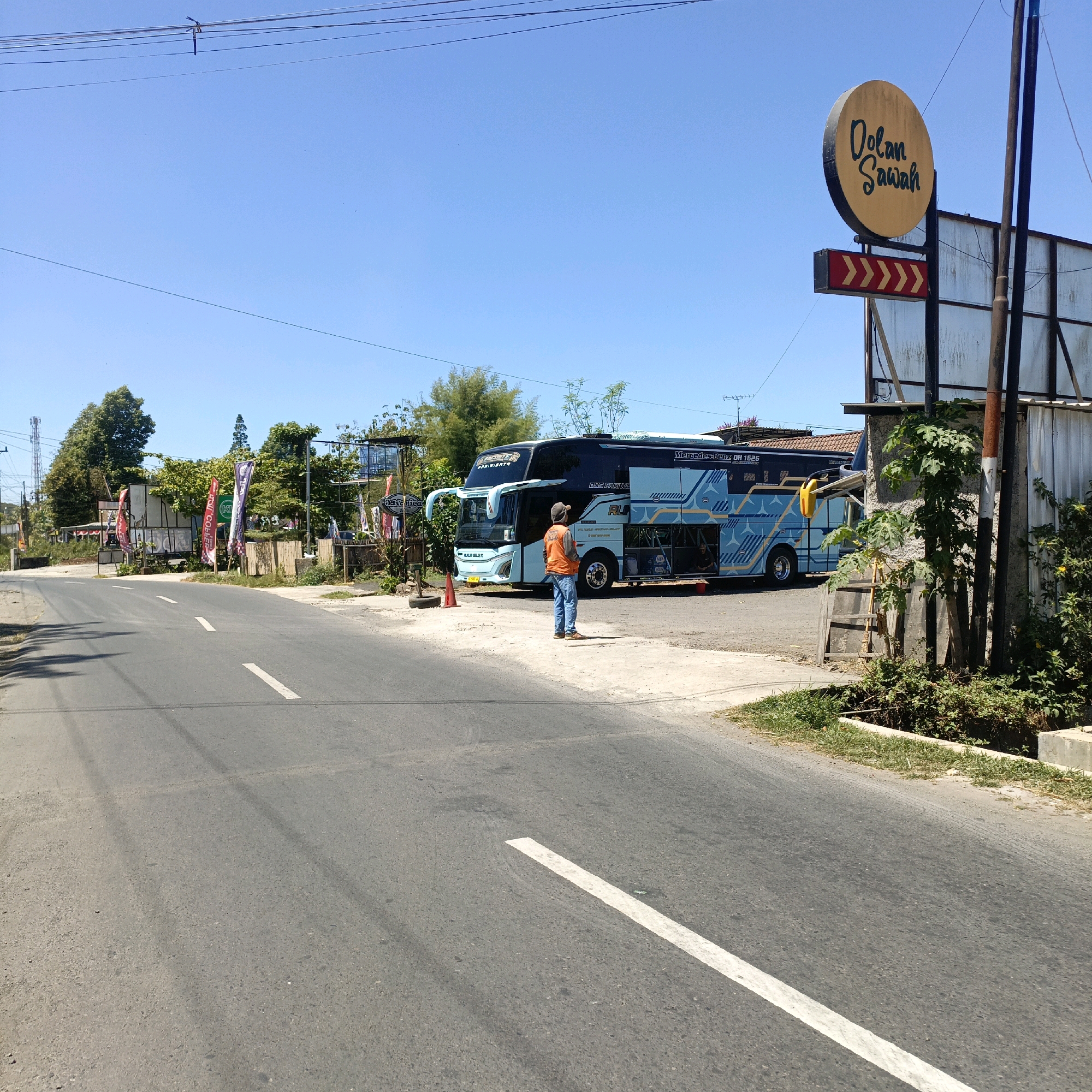 A light blue and black tourist bus is parked on the side of a paved road in an outdoor setting. The bus has "Mercedes-Benz OH 1626" written on it and features intricate blue and white patterned decals. A person wearing an orange vest, blue jeans, and a baseball cap is standing in front of the bus, facing away from the camera. The person appears to be holding something, possibly a phone, and is standing on a dirt and gravel area next to the road.
Behind the bus, there is a building with a white, corrugated metal roof and a light-colored facade. A circular sign with yellow lettering that reads "Dolan Sawah" is mounted on a pole, accompanied by a rectangular sign with red and yellow chevron patterns pointing to the right. Various colorful flags and banners are displayed along the roadside. Lush green trees and foliage are visible in the background. The sky is clear and blue, suggesting a sunny day. The road has a white dashed line marking the center. The environment appears to be semi-rural or on the outskirts of a town.
The specific location is identified as Jalan Gatot Subroto in Salatiga, Indonesia, a street known for its local eateries and tourist spots. The "Dolan Sawah" sign is a prominent marker for a place that offers leisure activities.