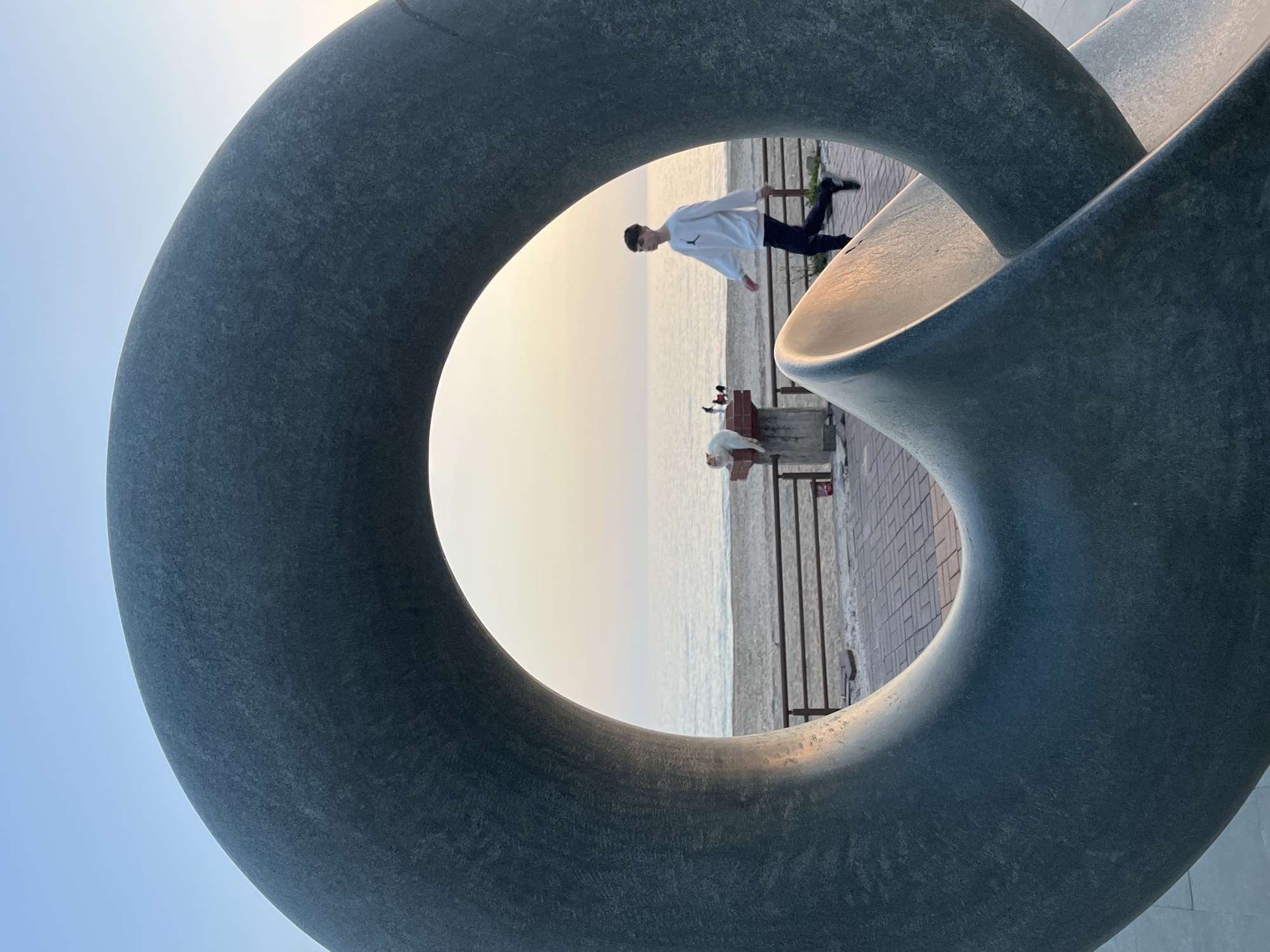 The image shows a large, gray, sculpted Mobius strip-like sculpture located in Alanya, Turkey. The sculpture is the focal point of the image, with its circular opening framing a view of the Mediterranean Sea, a section of the beach, and a nearby promenade.
Through the opening of the sculpture, a young man wearing a white shirt and dark pants is walking along the promenade. Near a low wall, there is a person lying on a bench. On the wall, there are also at least two cats. The sky is pale, suggesting either early morning or late afternoon. The promenade is paved with red bricks.