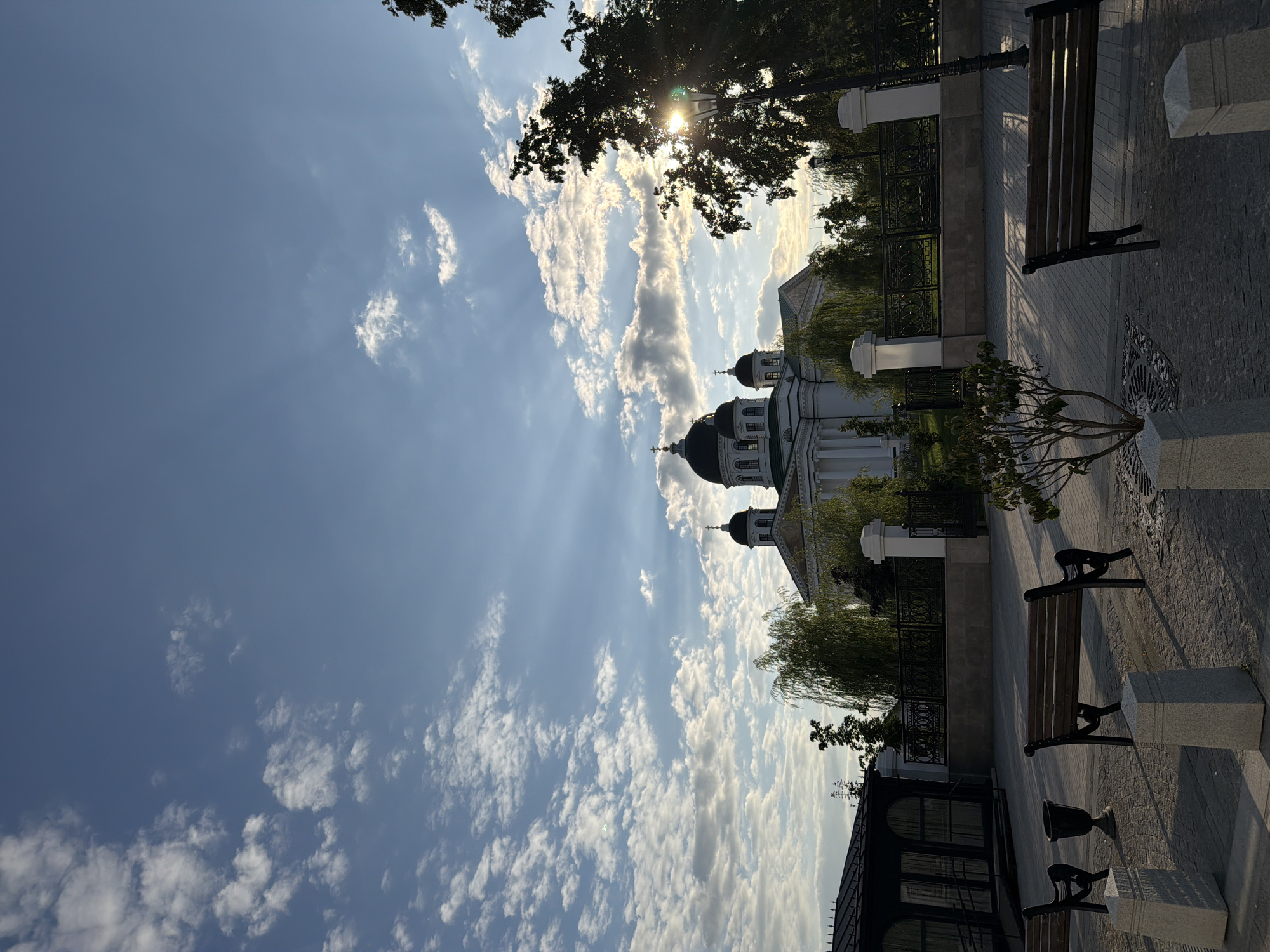 The image shows the Resurrection Cathedral in Arzamas, Russia. The cathedral is a large, white building with a green roof and multiple domes. It is set against a backdrop of a blue sky with white clouds. In the foreground, there are several benches and lampposts. The sun is setting, casting long shadows across the scene. The overall impression is one of peace and tranquility.
