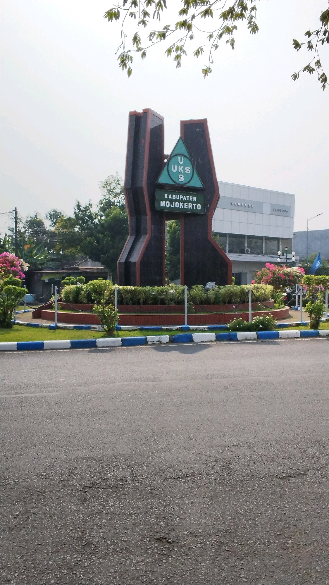 A monument with the text "UKS KABUPATEN MOJOKERTO" stands prominently in front of a modern building, possibly a car dealership, with "VINFAST" visible on its facade. The monument is dark colored with reddish-brown trim and features a green triangular emblem at its center. The area around the monument is landscaped with various shrubs and flowering plants, enclosed by a low wall with blue and white decorative trim. A paved road is visible in the foreground. The sky is overcast, and the overall scene suggests an outdoor public space in a developed area. The location is identified as Banjaragung, Indonesia.