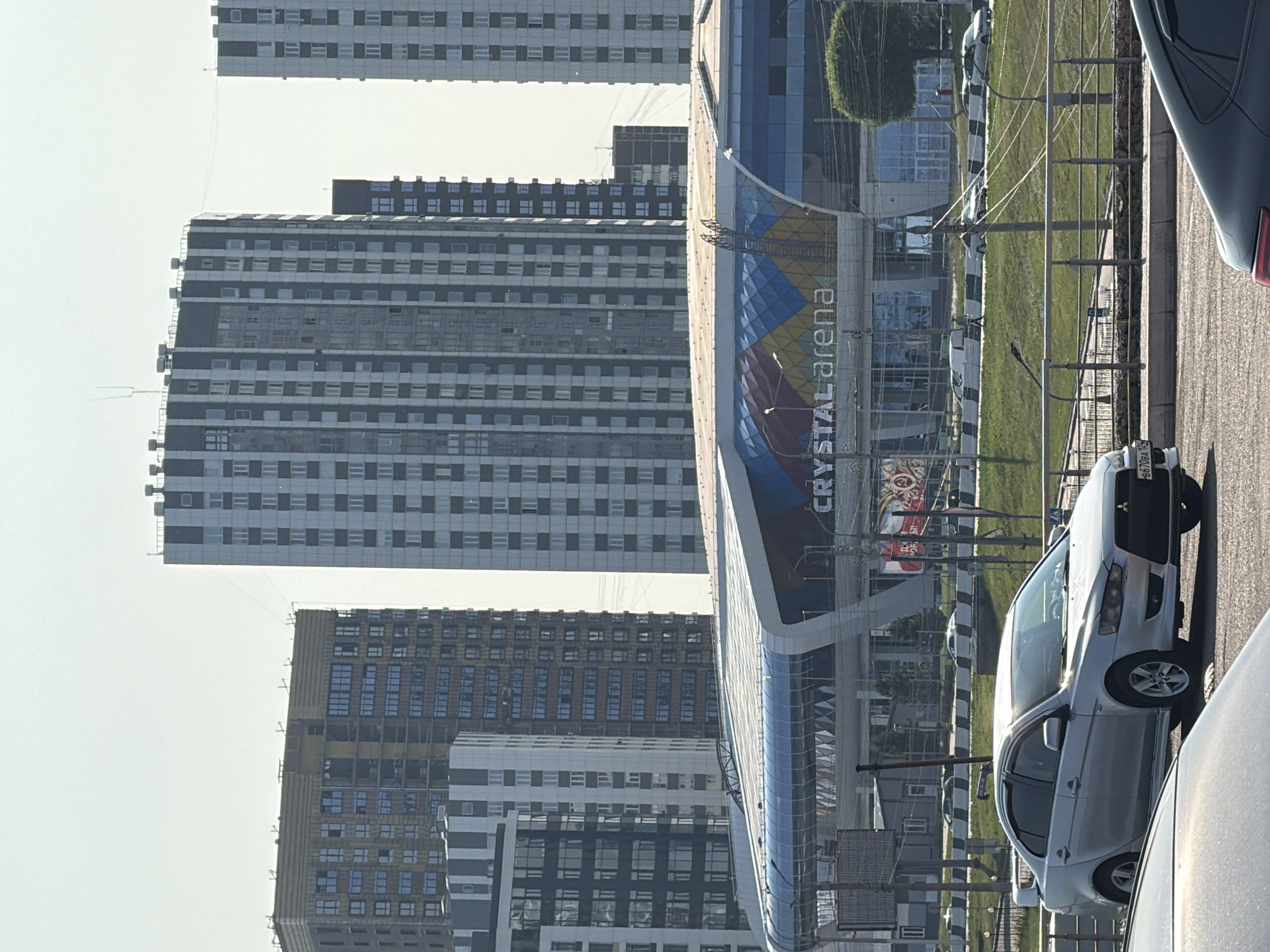 The image captures a street view in Krasnoyarsk, Russia, under clear, sunny skies, suggesting midday. The scene is dominated by a modern, curved building with a vibrant blue and yellow facade, clearly labeled "CRYSTAL arena" in white text. Adjacent to the arena are several tall, rectangular apartment buildings, their windows reflecting the bright light.
In the foreground, a silver Mitsubishi Lancer is parked on the right side of the road, its license plate partially visible. Another car, likely dark gray, is parked even closer to the viewer. A grassy area with a few trees and streetlights lies between the road and the arena. The overall atmosphere is one of urban development and everyday life, with no discernible emotions or specific events taking place beyond the typical activity of a city. The presence of multiple large residential buildings hints at a densely populated area.