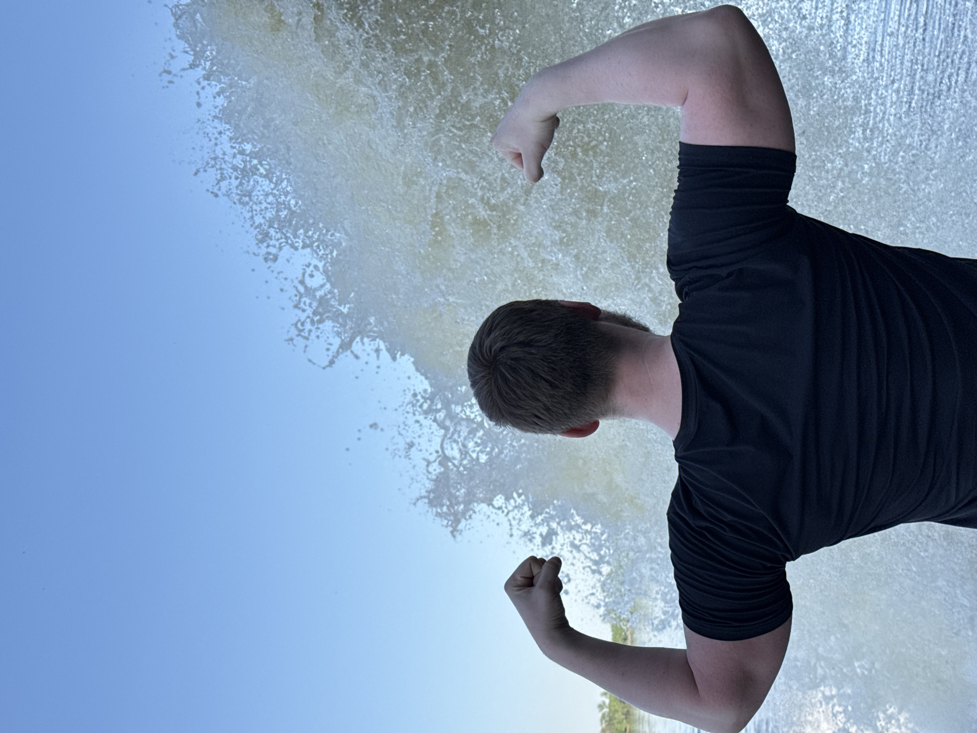 A man with short, light-brown hair, wearing a black t-shirt, is flexing his biceps. He stands in front of a large waterfall, and the sky is visible above it. The photo appears to have been taken from a lower angle, looking up at the man. The exact location in the city of Novyi, Russia, is not specified in the image.