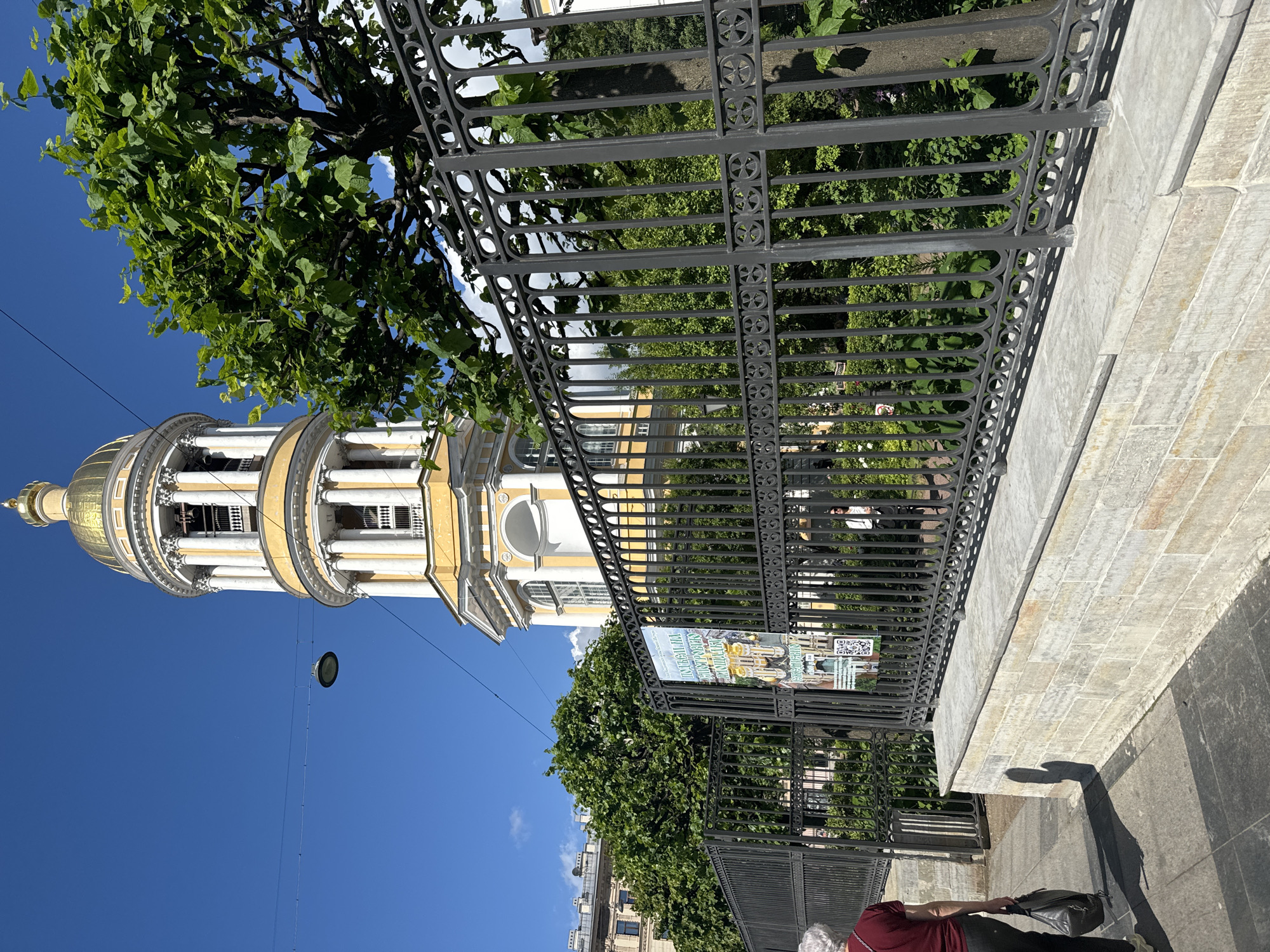 A yellow and white bell tower with a golden dome, topped by a small spire, is visible against a clear blue sky. The tower has multiple levels with columns and arched openings. In the foreground, a black wrought-iron fence with decorative circular patterns runs horizontally across the image. Behind the fence, lush green trees and foliage are abundant. A light-colored stone wall runs along the base of the fence. A person with gray hair wearing a maroon shirt and dark pants walks away from the camera to the left, carrying a black bag. Another person is partially visible through the fence in the background. A poster with images of buildings is attached to the fence. The scene appears to be outdoors, likely in an urban environment. The architectural style of the bell tower and the presence of the poster with Cyrillic text suggest the location is Saint Petersburg, Russia. The bell tower is part of the Vladimirsky Cathedral (also known as the Cathedral of the Vladimir Icon of the Mother of God), a prominent landmark in the city.