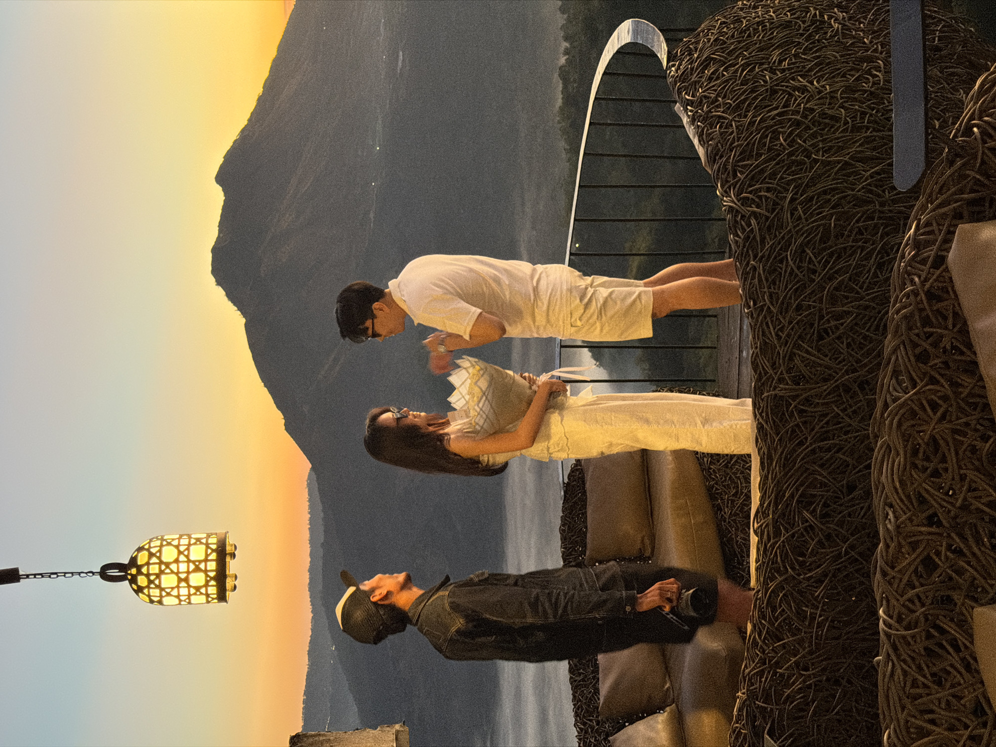 This image depicts a romantic scene at sunset overlooking Mount Batur in Kintamani, Indonesia. A man, dressed in a white polo shirt and shorts, holds a bouquet of flowers as he stands on a balcony, facing a woman in a white dress. The woman, wearing sunglasses, accepts the bouquet. To the left, another man in a black denim jacket and a baseball cap, holding a camera, observes the interaction. The balcony features woven seating and a railing, with the dramatic silhouette of Mount Batur and a misty valley in the background. A decorative lantern hangs from above, casting a warm glow. The sky is a gradient of orange, yellow, and blue, typical of a sunset.