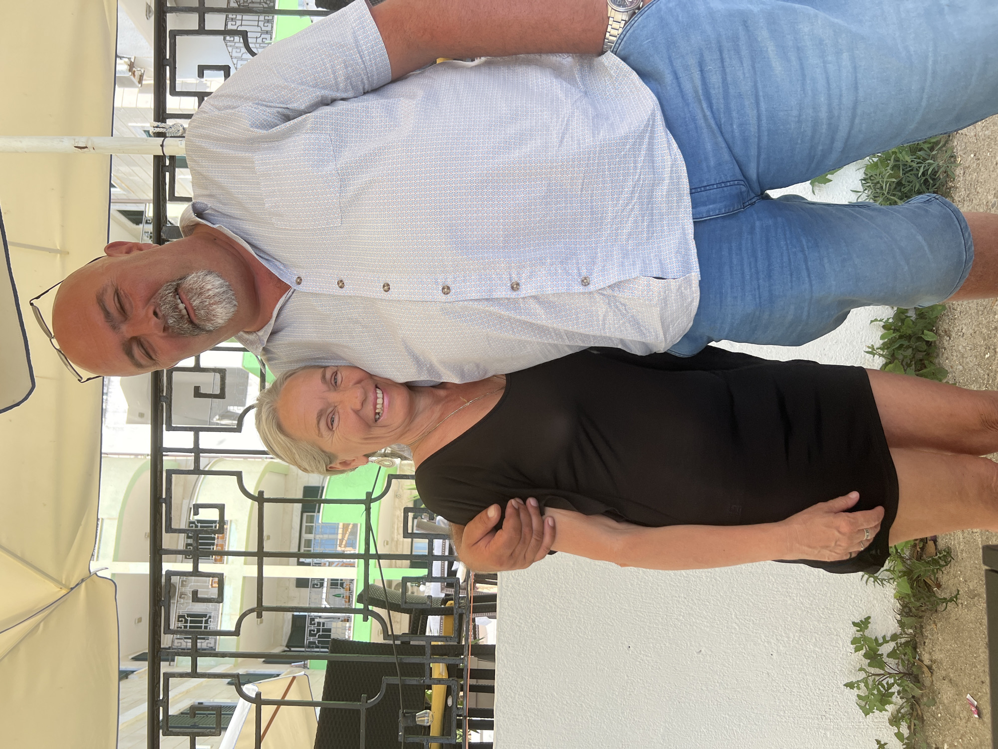 In Sveti Stefan, Montenegro, a middle-aged couple is pictured standing in front of a white wall and a decorative black metal fence. The man, with a bald head, grey goatee, and glasses perched on his head, wears a light blue patterned short-sleeved shirt and denim shorts. He has his right arm around the woman. The woman, with short, grey hair and a radiant smile, is dressed in a black short-sleeved dress and silver earrings. Her left hand rests on the man's arm. Behind them, a light-colored umbrella provides shade over the outdoor area, and a multi-story building with green balconies is visible through the fence. The ground is covered with gravel and patches of greenery.
