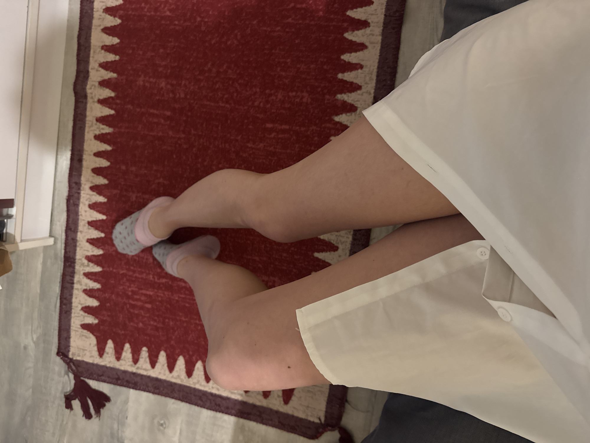 A high-angle shot depicts a person wearing pink socks and a white, button-down shirt sitting on a red rug. The rug is characterized by a geometric pattern with fringe at one end. The setting appears to be indoors, adjacent to a light-colored wall and a gray surface. The exact location is unspecified.