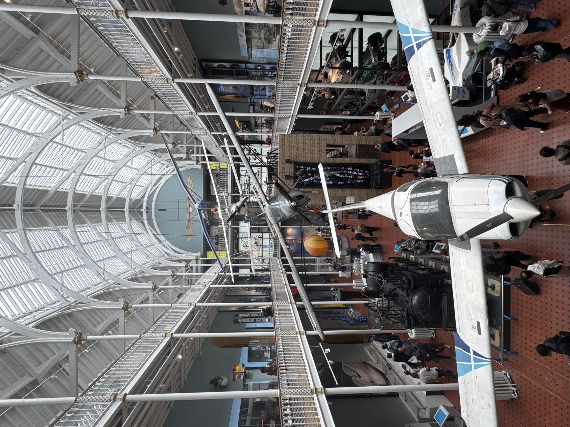 This is an eye-level shot of the Grand Hall at the National Museum of Scotland in the city of Edinburgh, United Kingdom. The hall is a vast, multi-story space with a glass-paneled, vaulted ceiling supported by white cast-iron columns and intricate latticework. Balconies with ornate white railings line the upper levels of the hall.
The main focal point of the image is a small, white propeller airplane suspended from the ceiling, with its wings extending across the frame. Below, the ground floor is a large open space with a patterned terracotta-tiled floor. Various exhibits are displayed throughout the hall. In the foreground, the nose and wing of another white airplane are visible. Towards the center of the image, a large black steam locomotive is on display. Other exhibits include what appear to be racing cars on elevated platforms and various historical artifacts and displays visible on the walls and within glass cases on the balconies.
There are numerous people populating the museum, walking around the exhibits and on the balconies. They are dressed in casual clothing, and their actions suggest they are visitors exploring the museum. Some are looking at the exhibits, while others are walking or talking amongst themselves. The lighting in the hall is bright, coming from the overhead skylights and artificial lighting. The overall atmosphere is one of a busy public museum.