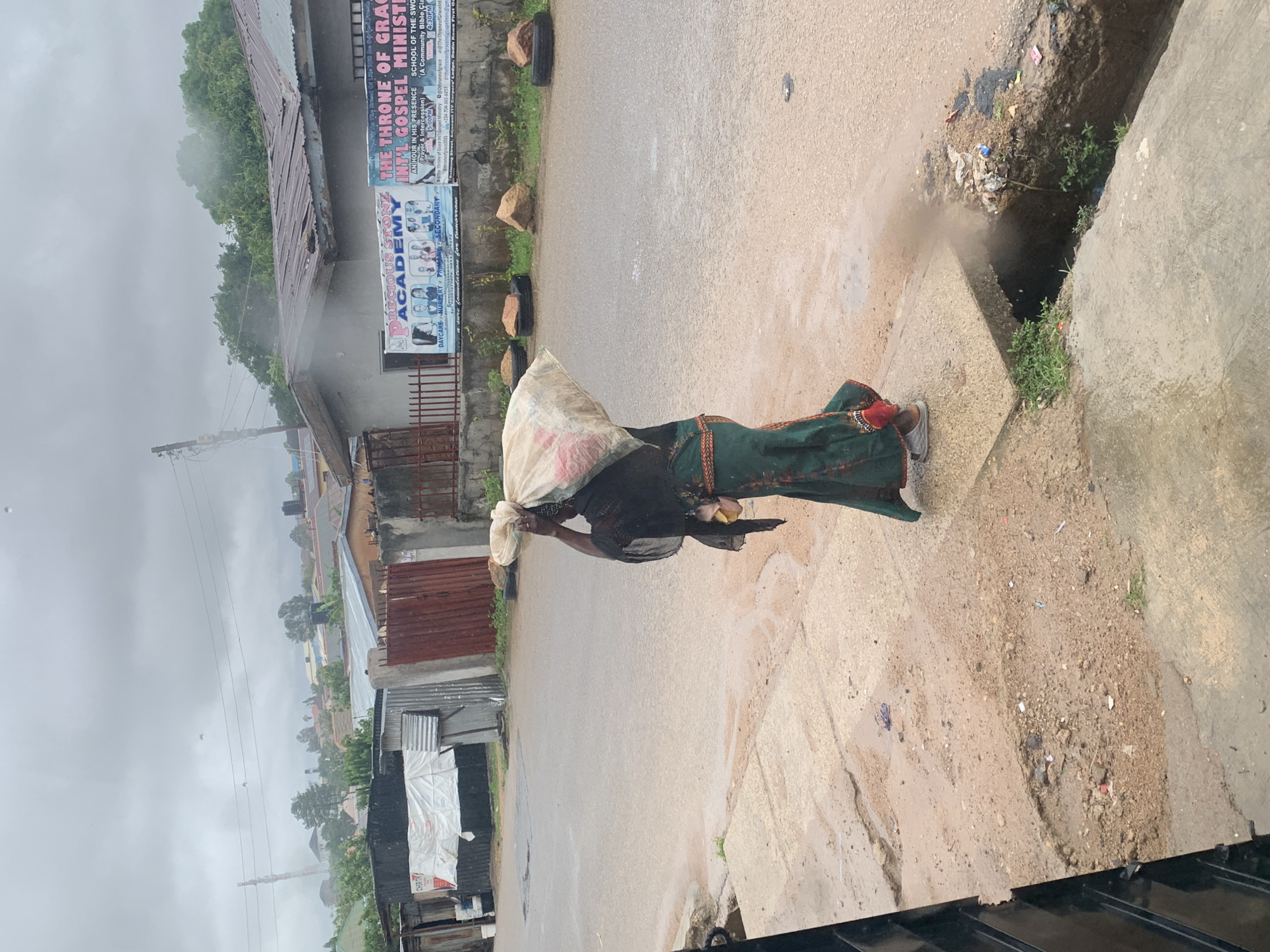 This image, taken in Jos, Nigeria, captures an overcast day scene on a street with a person walking along a concrete path next to a dirt-and-concrete drain. The main subject is an individual, likely female, seen from the back, wearing a dark top, a patterned green and red skirt, and light-colored sandals. They are carrying a large, light-colored sack or bag, possibly white, on their head, and appear to be holding a small item in their right hand.
The street itself appears to be paved with asphalt or a similar material and shows signs of recent wetness. To the right of the walking path, there is an open ditch or drain containing dirt, some small green plants, and visible litter, including plastic debris.
In the background, there are various buildings with corrugated metal roofs, typical of the region. Advertising banners are prominently displayed on one building. One banner advertises "PRECIOUS STONE ACADEMY" offering "DAYCARE NURSERY PRIMARY SECONDARY," with a slogan "a sure foundation for tomorrow." Another banner announces "THE THRONE OF GRACE INT'L GOSPEL MINISTRY" and "SCHOOL OF THE SWORD (A Community Bible Class)" with service times like "THUR 4:30PM" and "6:00PM," and contact information including phone numbers and social media handles like "@Othroneofgrace."
The sky is heavily overcast, suggesting either an impending rain, ongoing light rain, or recent rainfall, contributing to a muted light and a slightly misty appearance in the distance. Utility poles and power lines are visible, extending into the hazy background. Scattered debris and rocks are present along the roadside, with old tires used as informal barriers or decorations near the buildings. The word "CHERIT" is visible on the sack carried by the person, possibly indicating a brand or material.