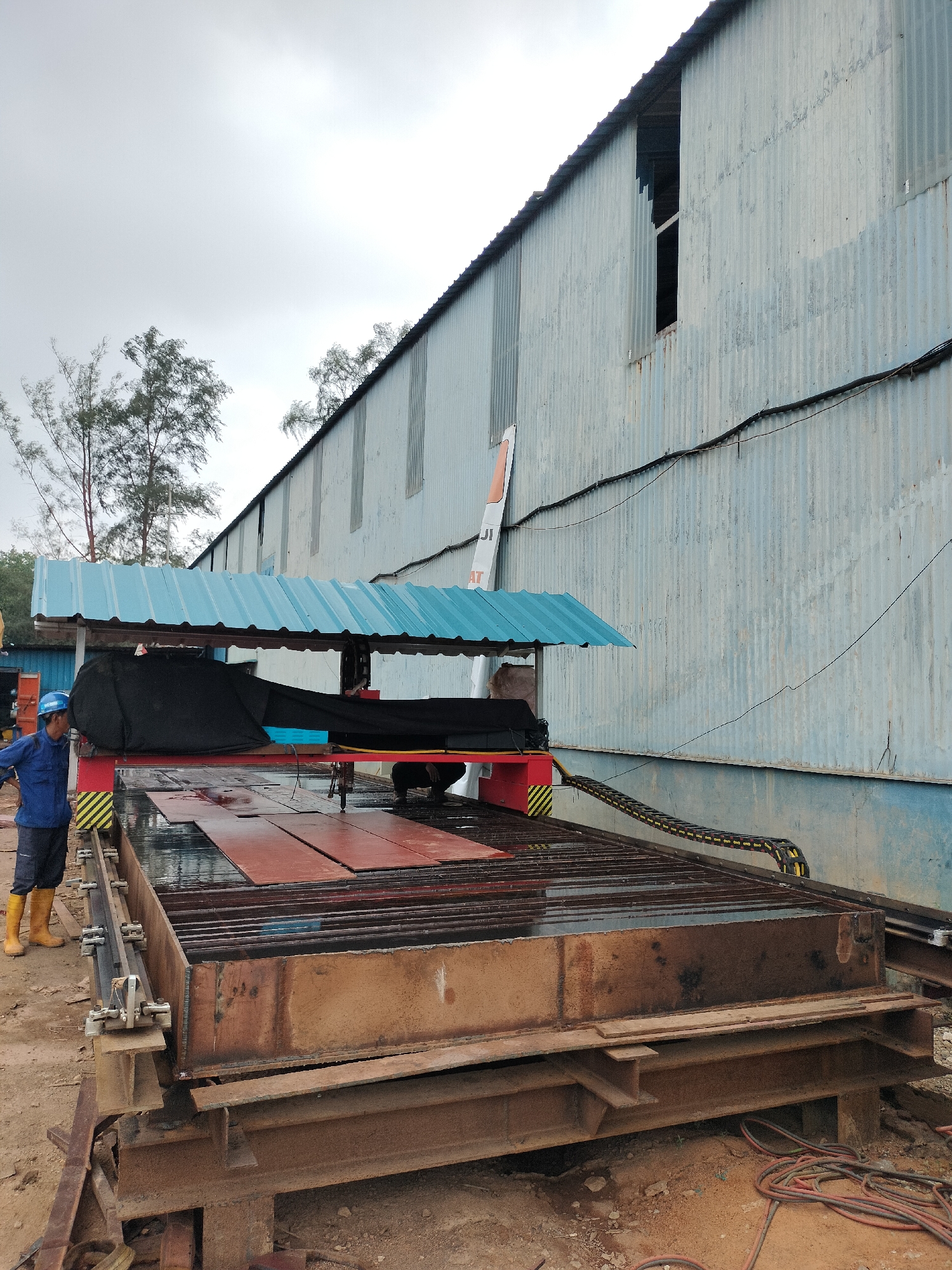 A person wearing a blue uniform and yellow boots stands next to a large metal cutting machine in what appears to be an industrial area in Batam, Indonesia. The machine has a red and black cutting head assembly and is positioned over a bed of metal plates. The background features a corrugated metal building with a blue trim and some trees. The sky is overcast. The scene suggests an active worksite where metal fabrication is taking place.