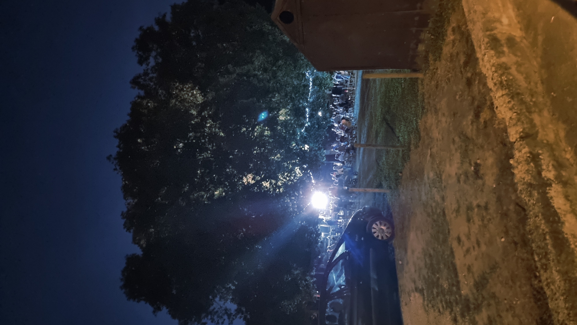 In Sant Joan Les Fonts, Spain, a night scene unfolds with a large tree dominating the background, its leaves dimly illuminated by scattered lights. A group of people are gathered in the mid-ground, seated on chairs and appearing to watch something. A bright light source is visible in the center of the gathering, casting rays through the tree. To the left, a dark-colored car is parked. To the right, a wooden structure with a dark roof stands. The ground in the foreground consists of dirt and patches of grass. String lights are visible suspended from the branches of the tree, adding to the ambiance.
