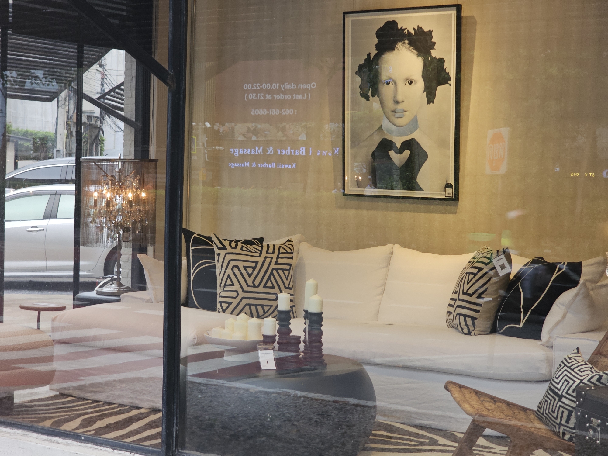 The image shows a storefront window display in Bangkok, Thailand. The display features a modern living room setting with a white sectional sofa, decorative pillows with geometric patterns, and a round coffee table with candles. A framed abstract portrait of a woman hangs on the wall behind the sofa. Reflected in the window are street scenes, including a silver car and building facades. Text on the window, visible as a reflection, indicates business hours and services, possibly for a salon or spa, mentioning "Kwanruedee Barber & Massage." The overall impression is one of contemporary interior design displayed in a commercial setting within a bustling city environment.