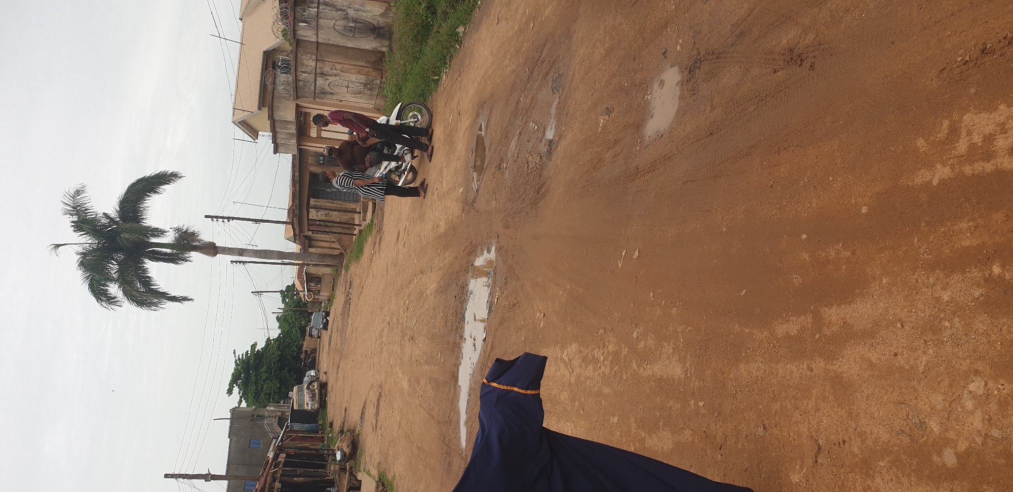 In Sabon Tasha, Nigeria, an overcast sky looms over a dirt road filled with tire tracks and puddles. To the left, a dark blue garment with an orange trim hangs in the foreground, obscuring part of the scene. Further down the road, a tall palm tree stands prominently against the sky. On the right side of the road, three individuals are gathered around a white motorcycle. One person, wearing a black and white striped shirt and black pants, stands on the left, facing the motorcycle. Another person, dressed in a brown shirt and dark pants, sits on the motorcycle. A third individual, in a maroon shirt and black pants, stands on the right, facing the person on the motorcycle. Buildings with corrugated metal roofs and concrete walls line both sides of the street, some appearing to be shops with open fronts. A few vehicles, including what appears to be a pickup truck and a car, are parked or driving in the background. The overall impression is of a typical, somewhat unkempt, urban street scene.