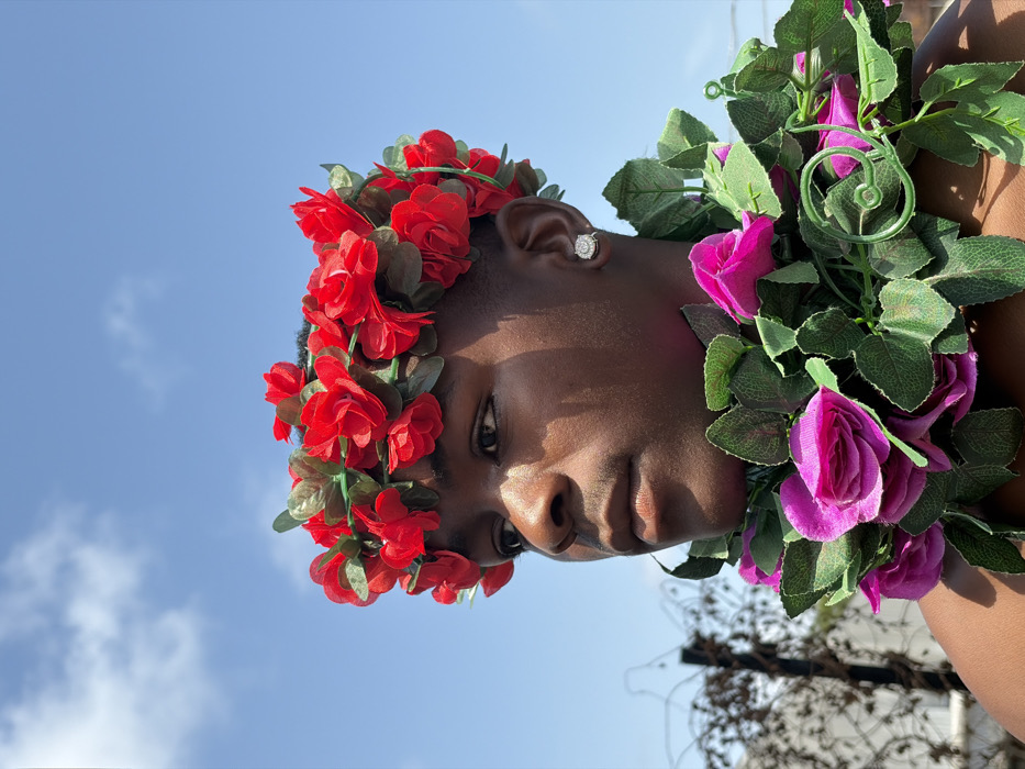 A dark-skinned male individual is depicted outdoors in Igboefon, Nigeria. He is positioned facing slightly to the left, with his gaze directed off-camera. A crown of artificial red roses and green leaves rests on his head. An artificial garland of magenta roses and green leaves is draped around his neck and shoulders. A round, light-colored earring is present in his visible right earlobe. The scene is illuminated by bright, direct sunlight, casting distinct shadows on the subject's face. The background features a clear blue sky with scattered white clouds above. To the left, a blurred dark fence post with visible barbed wire elements and indistinct light-colored structures are present. No other individuals or explicit actions are observed.