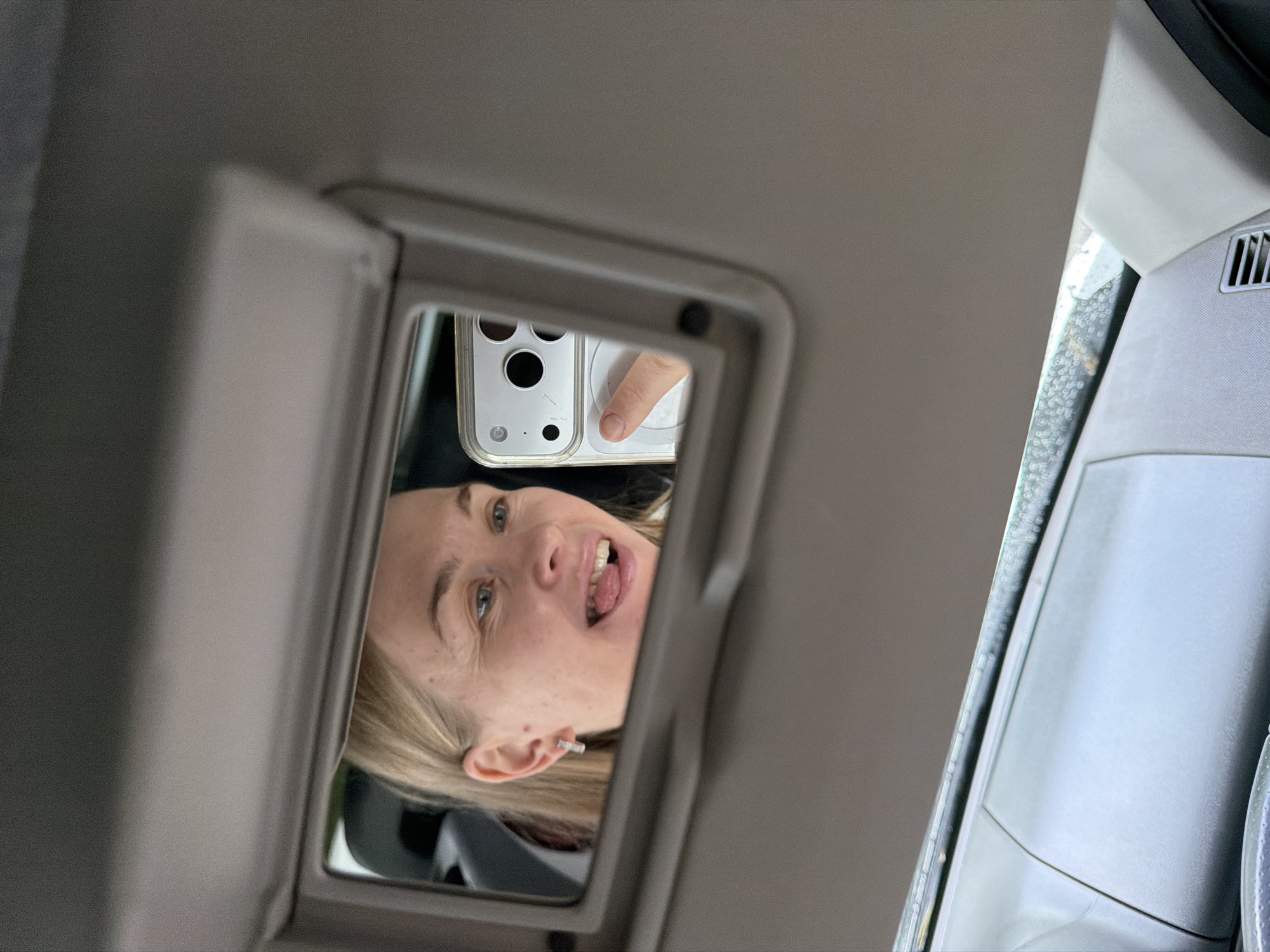 The image is a selfie taken in a car's visor mirror by a woman with blonde hair. She is sticking her tongue out and wearing a small, rectangular earring. She is holding a white smartphone to take the picture. The background of the car interior is visible, along with rain on the windshield. The image does not contain any information that would indicate its location in the city of Khabarovsk, Russia.