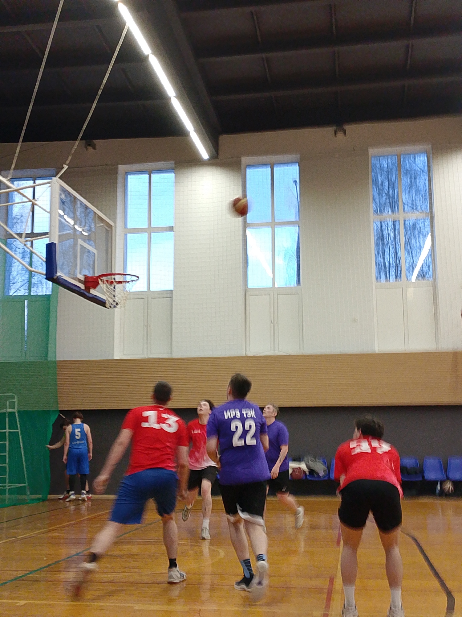 A basketball game is in progress inside a gymnasium in Izhevsk, Russia. Several male players are in motion on a polished wooden court, wearing jerseys with numbers: 5 in blue, 13 in red, 22 in purple, and another in red (number not visible). A basketball is airborne between the players and a basketball hoop with a net. The gym has a white-tiled wall with three large windows revealing leafless trees outside. A light fixture illuminates the scene from the ceiling. There is a green divider on the left side. In the background, blue chairs are visible.